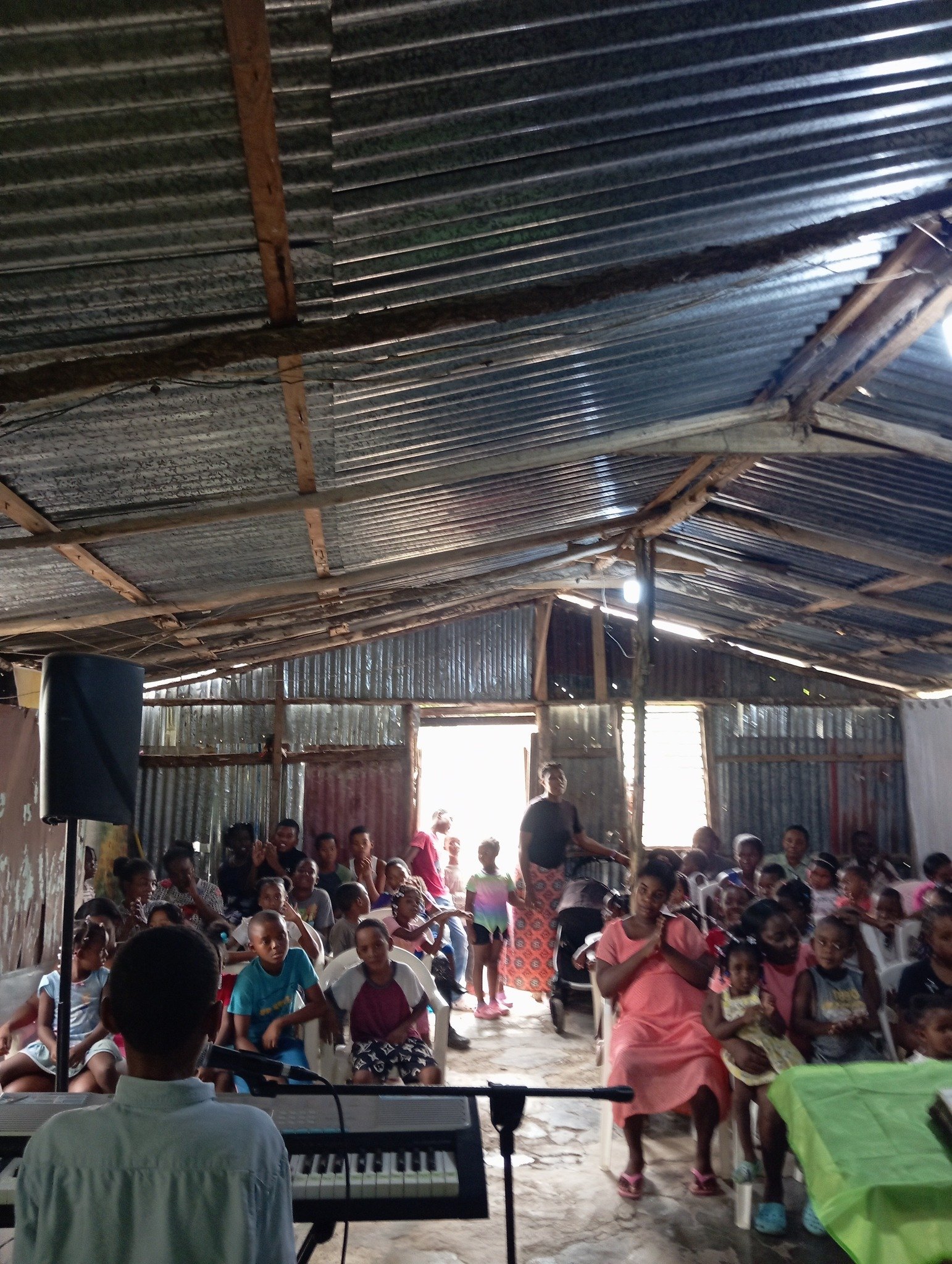 A group of children and adults sitting inside a rustic, metal-roofed building with open windows, with a person playing a keyboard at the front.
