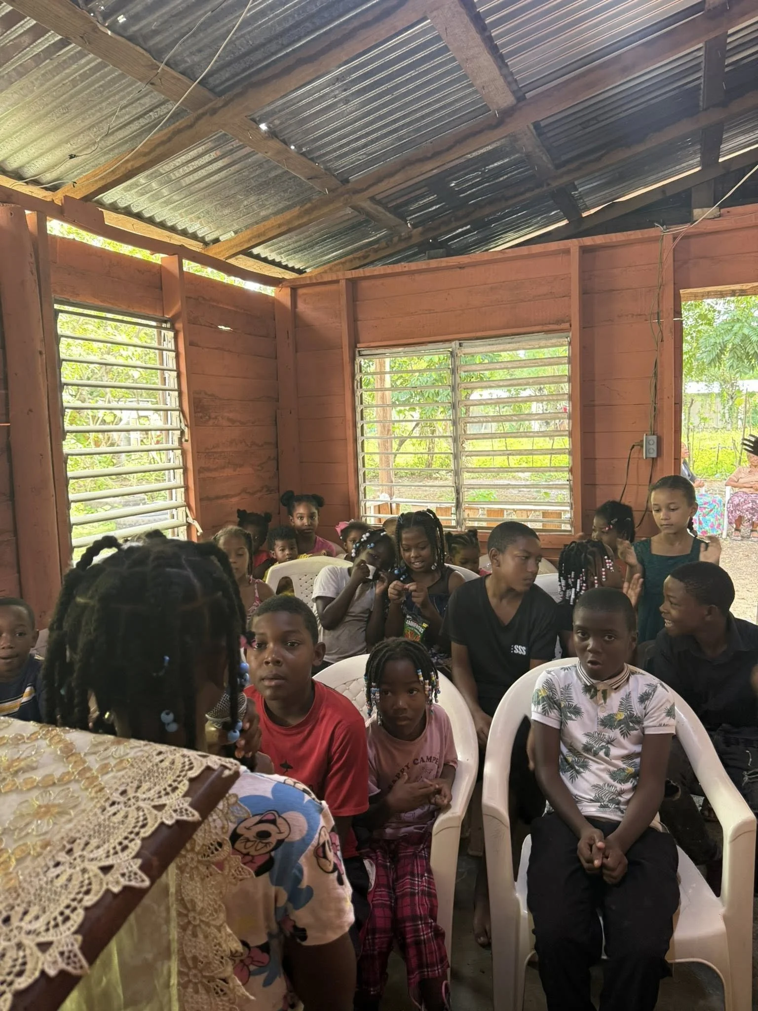 Group of children sitting and standing inside a wooden structure with open windows, listening to a person speaking at the front.
