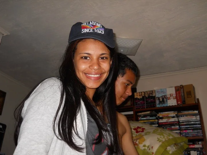 Smiling woman wearing a dark baseball cap with rainbow-colored embroidery, standing indoors with a young man in the background, bookshelves, and a floral pillow.