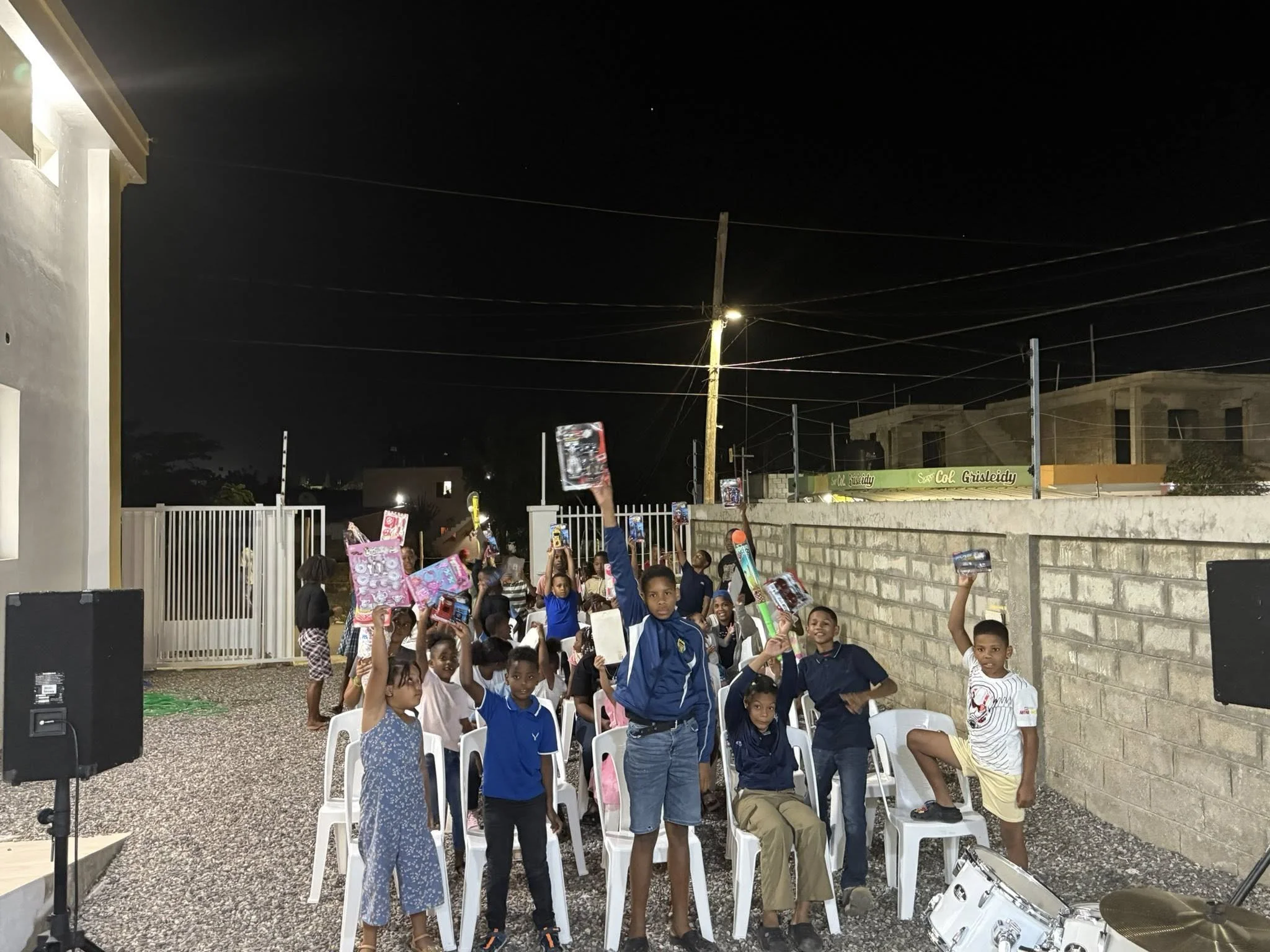 Children at night outdoors holding and raising toys, some sitting on white chairs, with a wall and fence behind them, and a party atmosphere.
