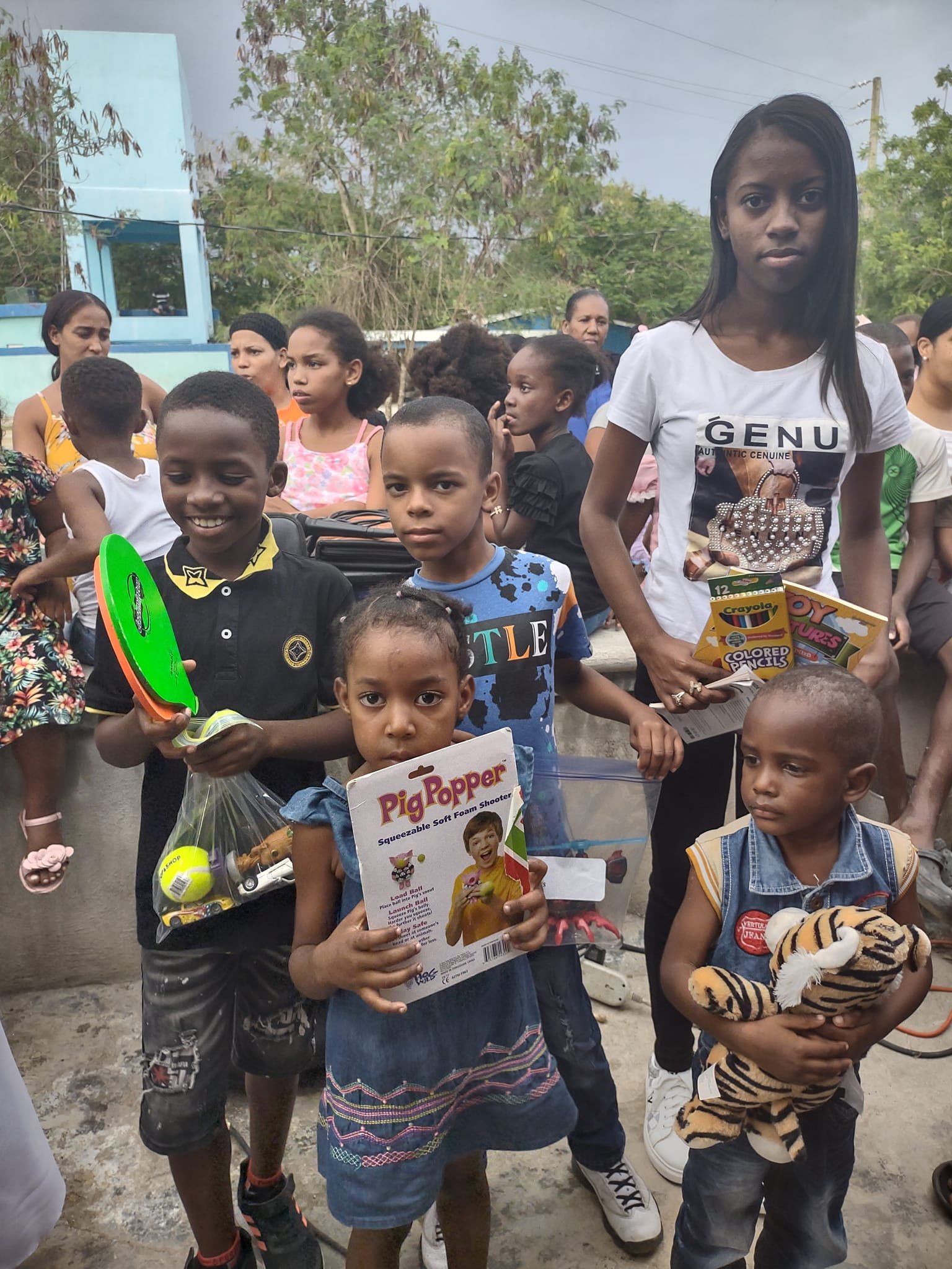 Group of children and women gathered outdoors, some holding toys and colorful packages, in a casual setting with trees and a blue building in the background.