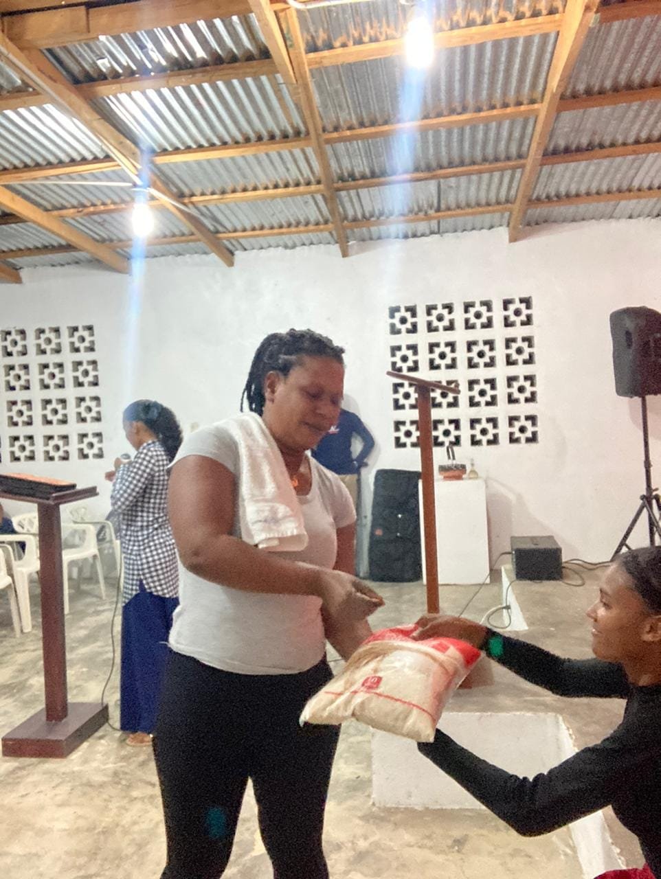 A woman receiving a bag of rice from another woman seated on a white chair in a community hall with white walls, wooden ceiling, and decorative concrete block windows.