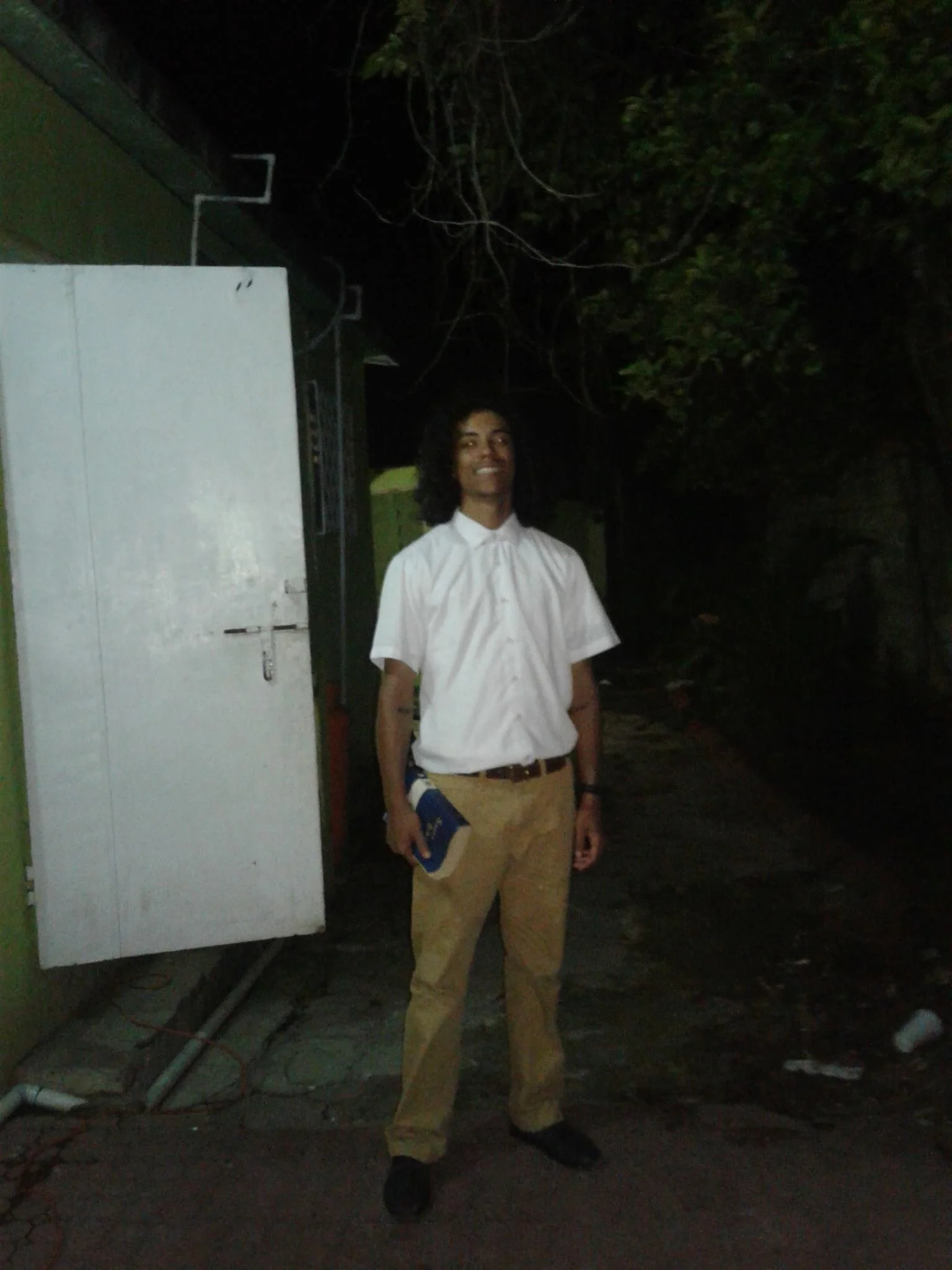 A person with long curly hair, wearing a white button-up shirt and tan pants, standing outdoors at night, holding a book or notebook in their left hand, smiling, near a green building with an electrical box and wires.