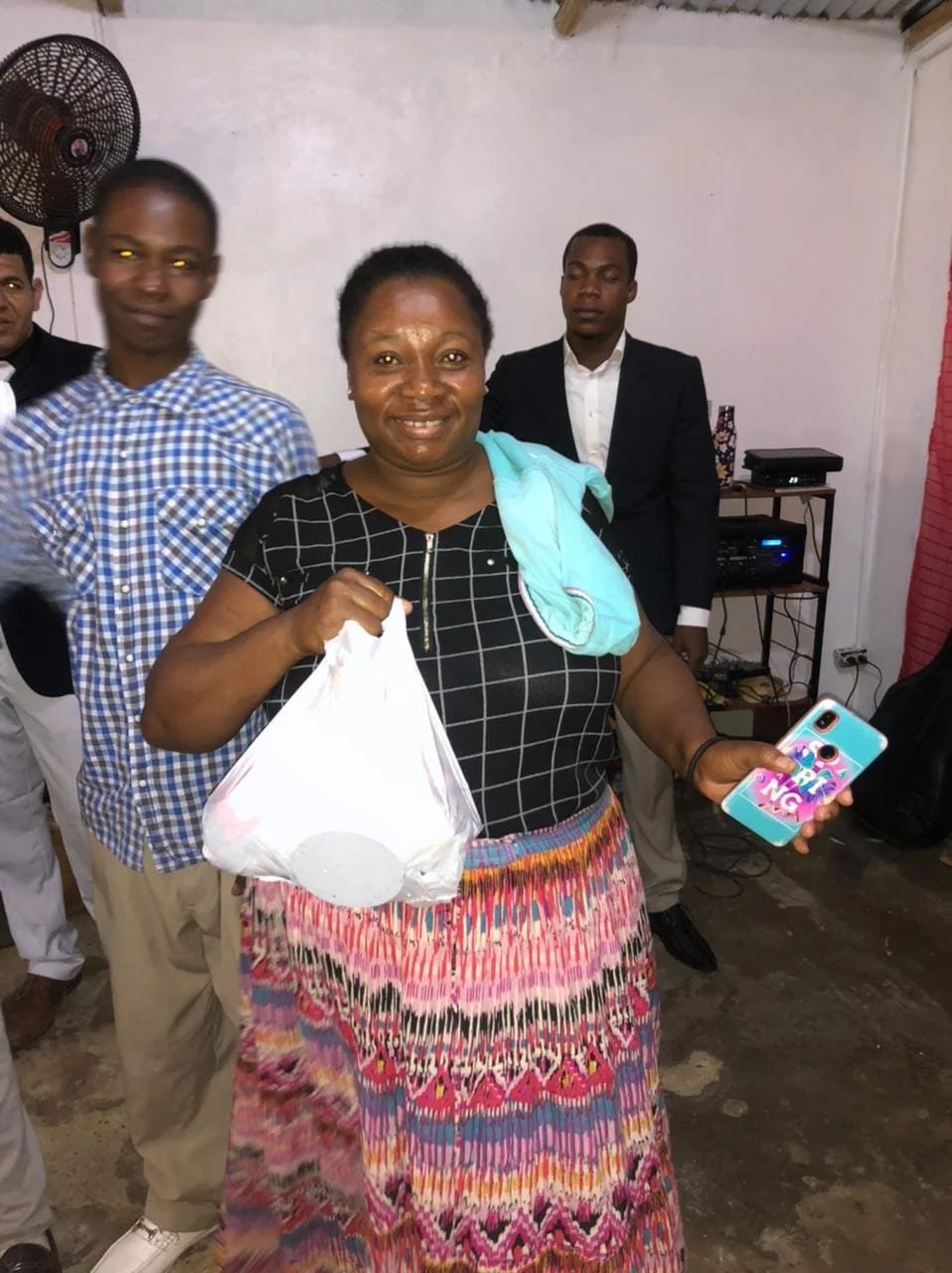 A smiling woman holding a plastic bag and a colorful phone in a room with men in the background.