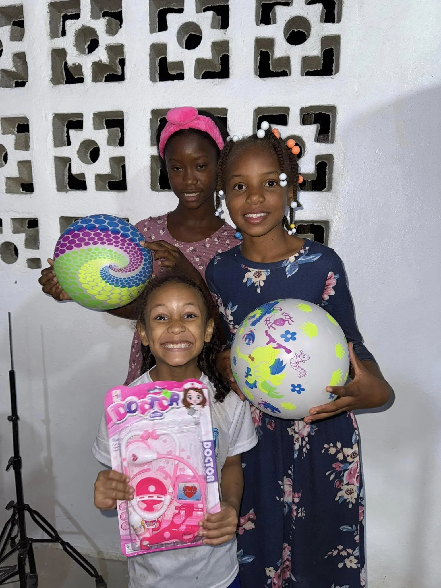 Three young girls holding balloons and toy stethoscope. One girl in the front has a big smile, holding a pink toy stethoscope, while the other two girls behind hold colorful balloons, all smiling at the camera.