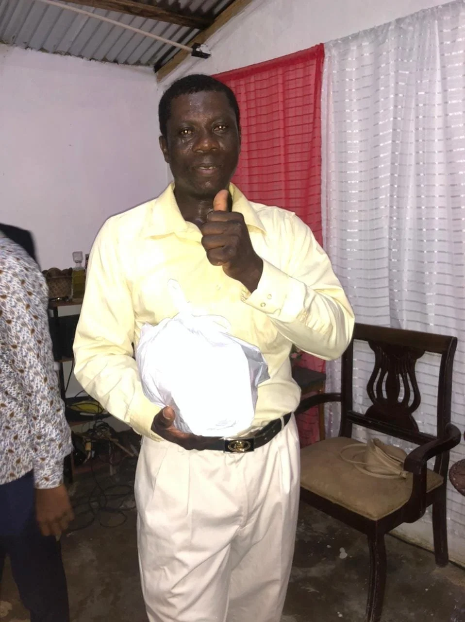 A man in a yellow shirt giving a thumbs-up, holding a wrapped gift, in an indoor setting with red and white curtains in the background.
