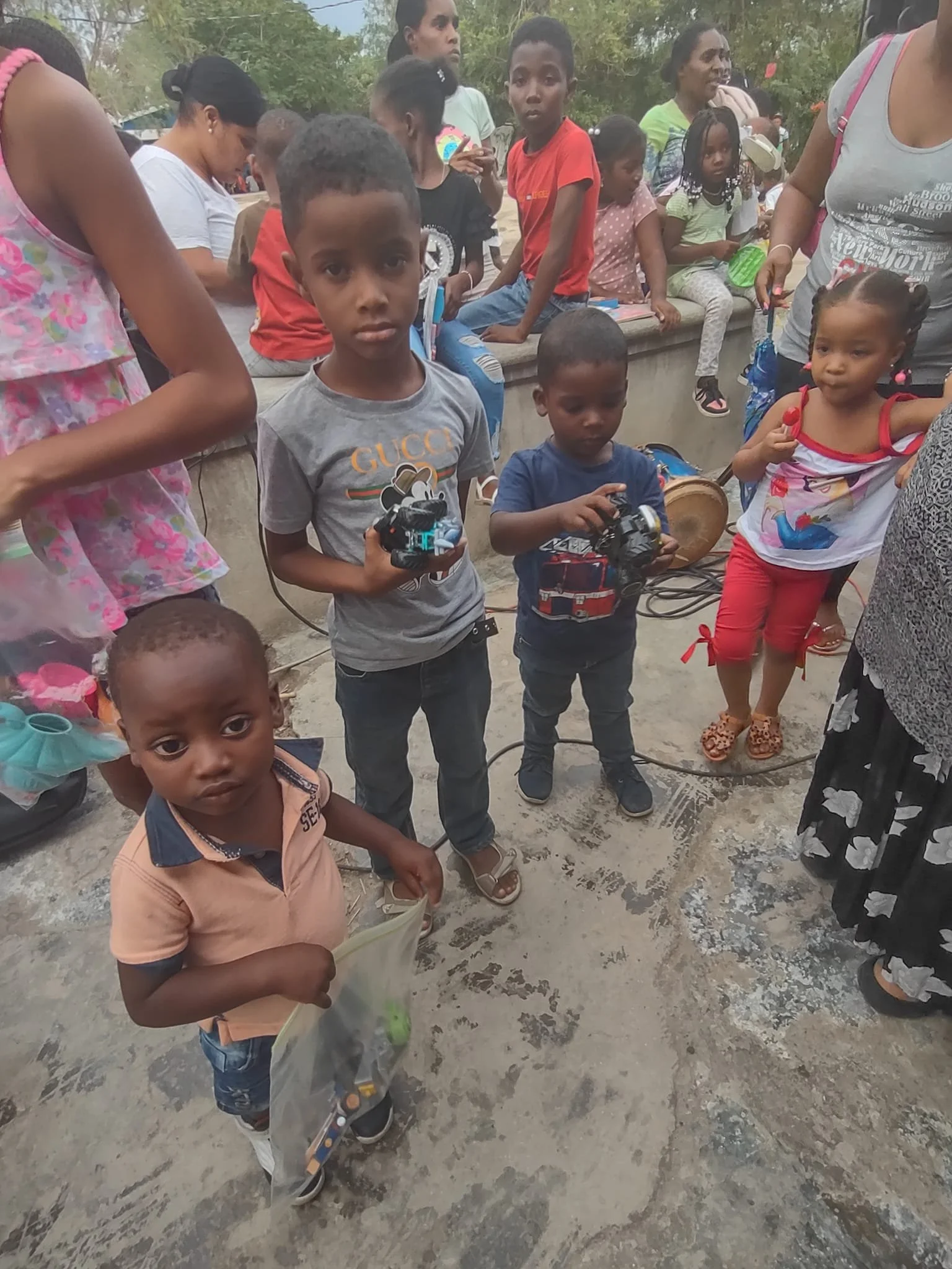 Group of young children and adults gathered outdoors, with some children holding cameras. The children appear curious and serious, standing on a concrete surface with greenery in the background.