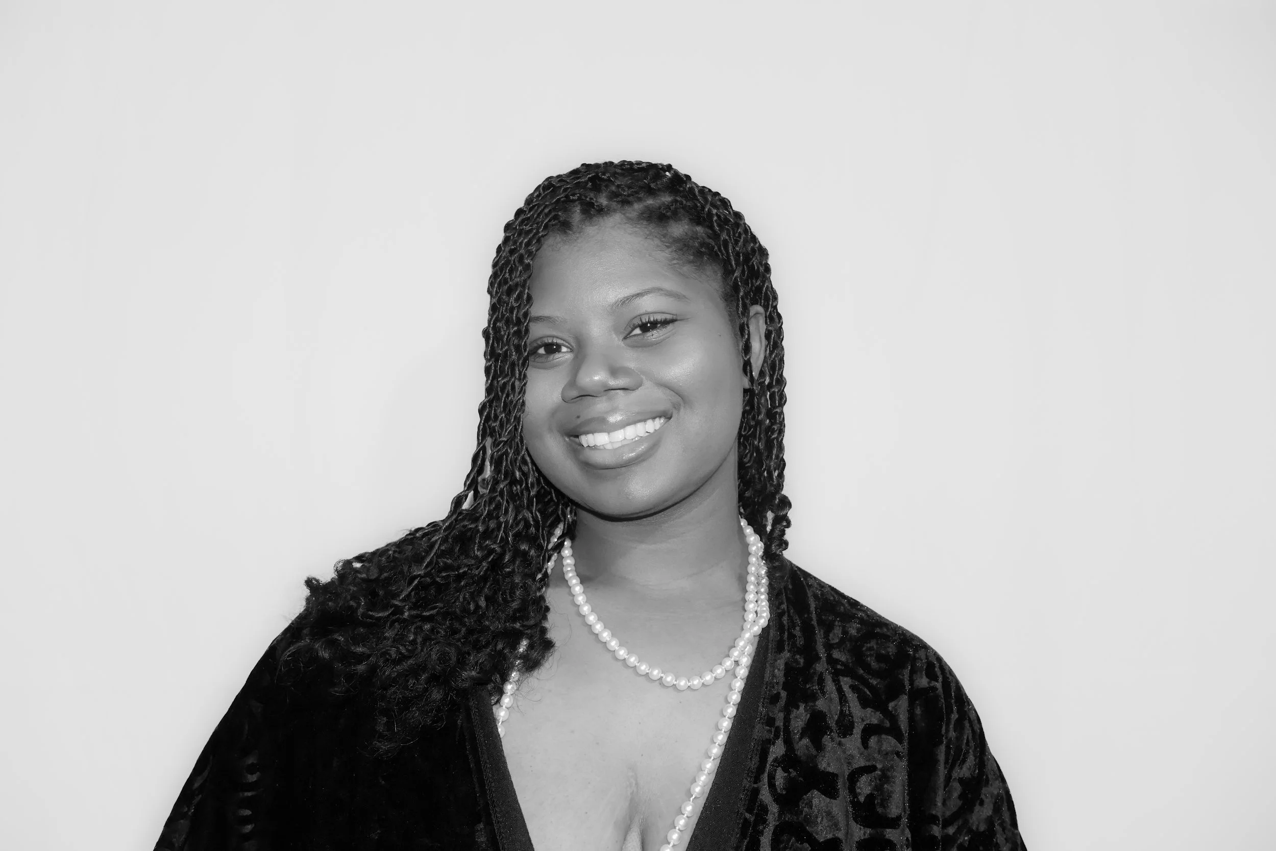 A woman with braided hair smiling, wearing a pearl necklace and a dark patterned top, against a plain light background.