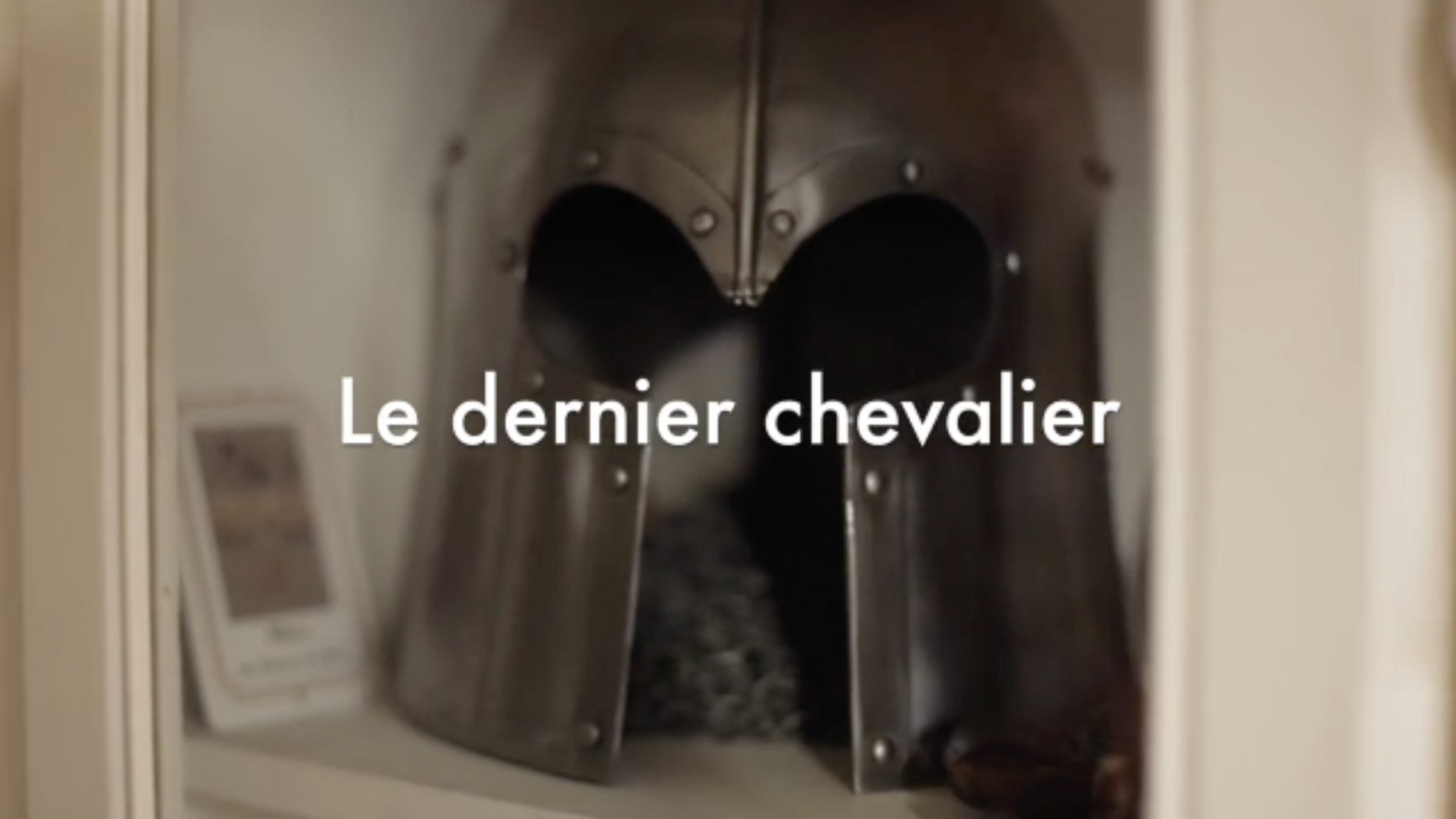 Une armure en métal brillante de style chevalier avec un casque intégral, vue de face dans une vitrine avec une légende en français 'Le dernier chevalier'.