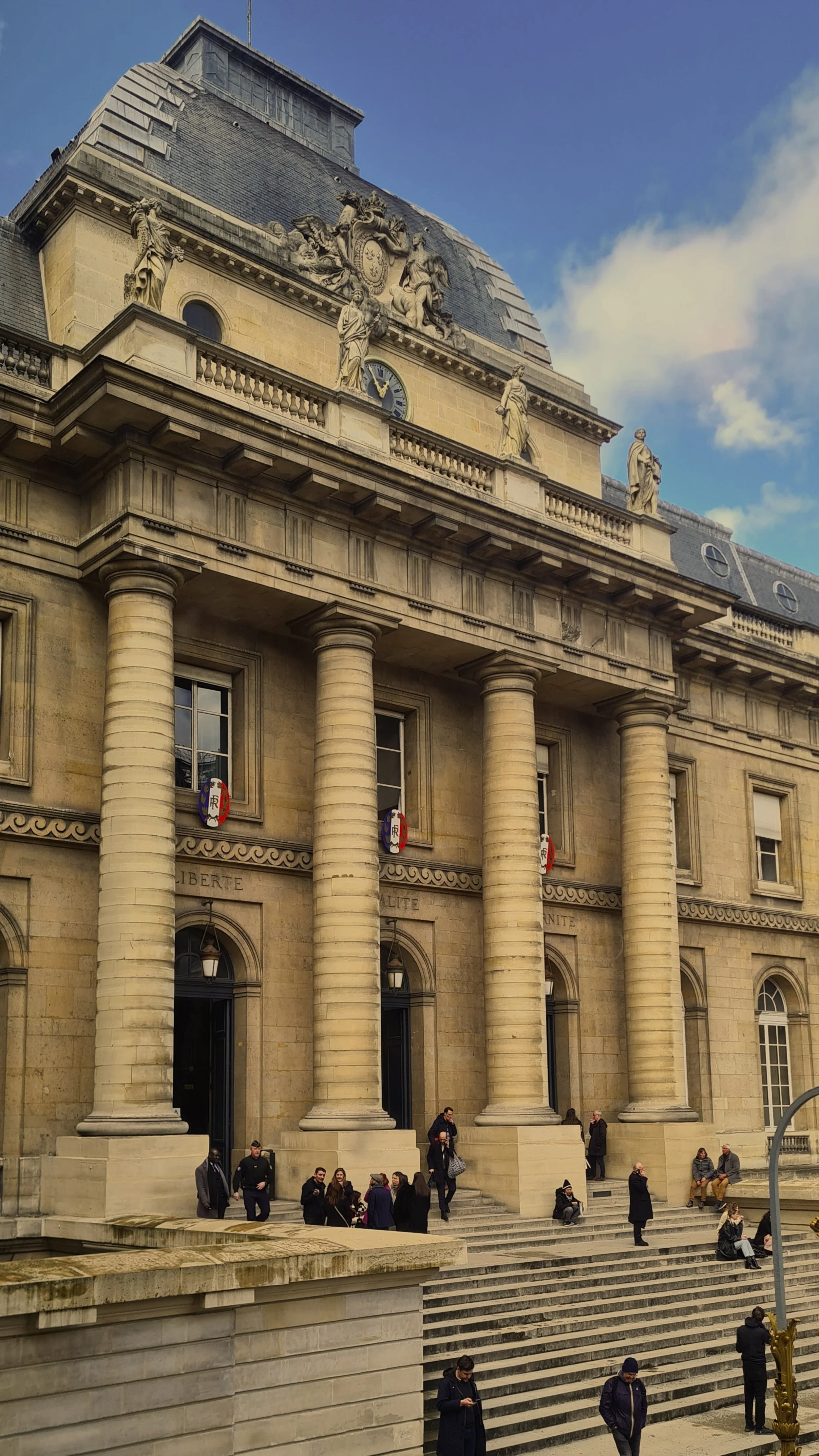 Une façade d'un bâtiment traditionnel avec des colonnes, des statues et un drapeau français, avec des personnes assises ou discutant sur les marches.