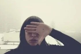 Une personne avec les cheveux longs et noirs, portant un sweat noir, cache une partie de son visage avec sa main dans un environnement intérieur minimaliste.