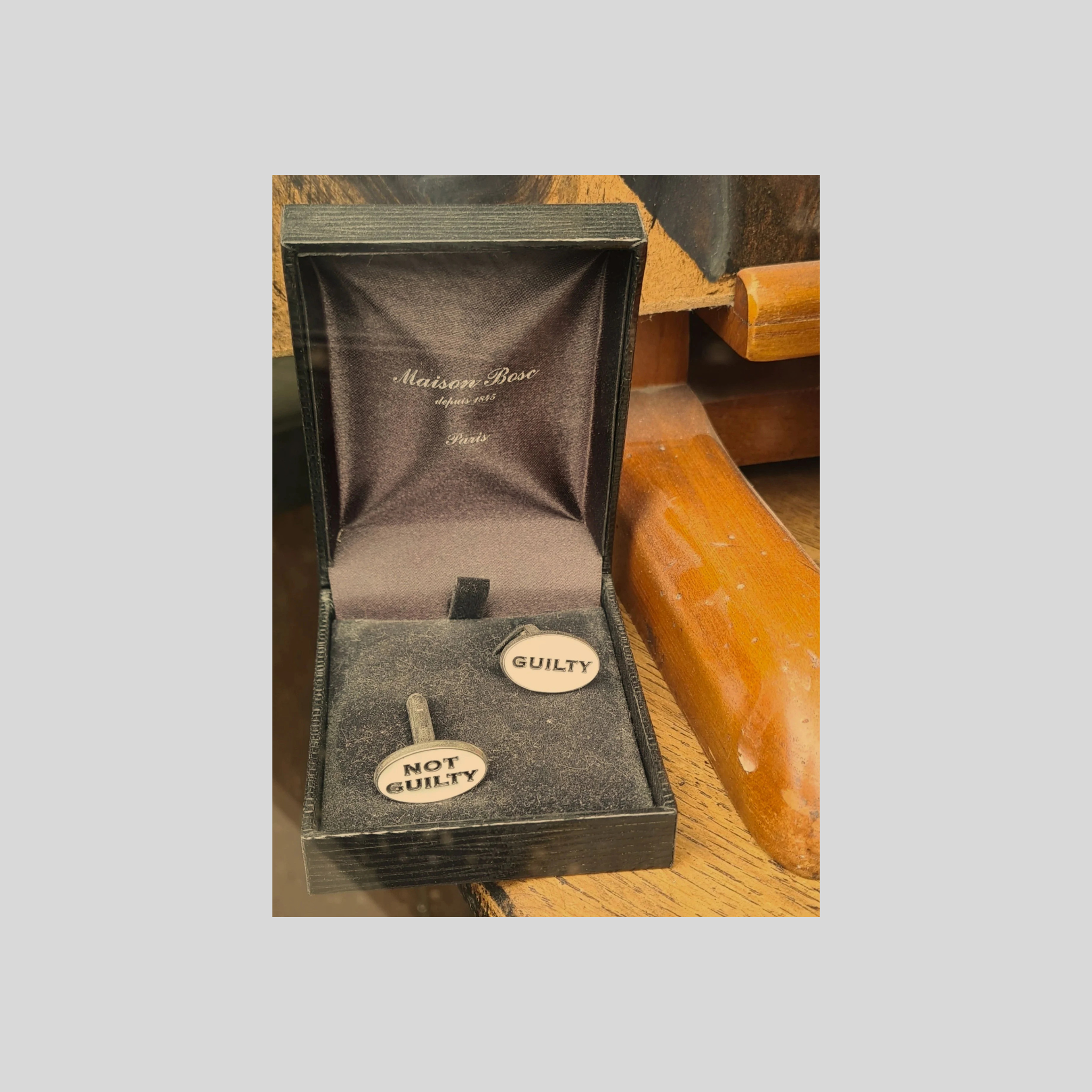 Une boîte à souvenirs avec deux pin’s, l’un marqué 'GUILTY' et l’autre 'NOT GUILTY', posée sur une surface en bois, avec un fond de mobilier en bois.