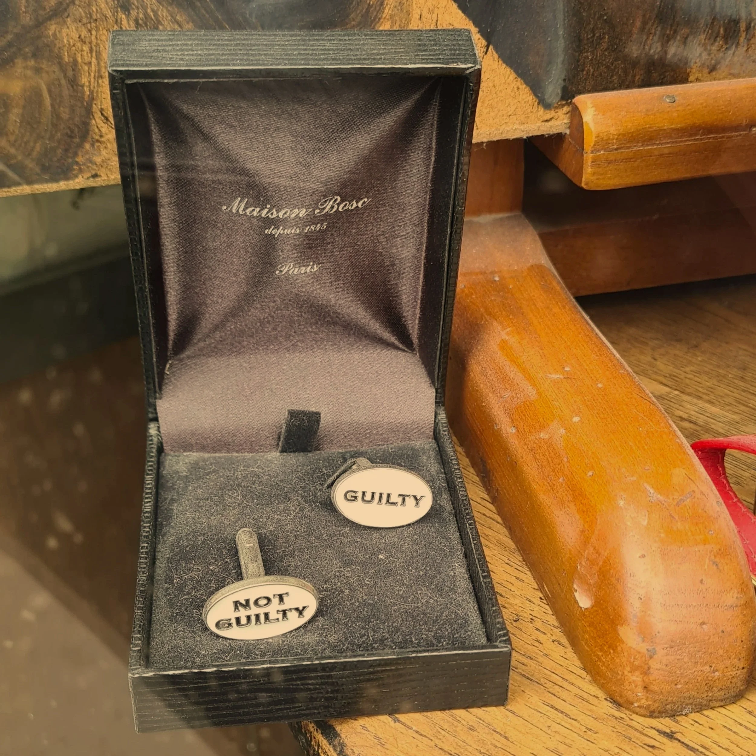 Boîte ouverte contenant deux boutons de mariée avec inscriptions 'GUILTY' et 'NOT GUILTY' sur un fond en velours, la boîte porte l'inscription 'Maison Bosc, since 1815, Paris' à l'intérieur du couvercle.