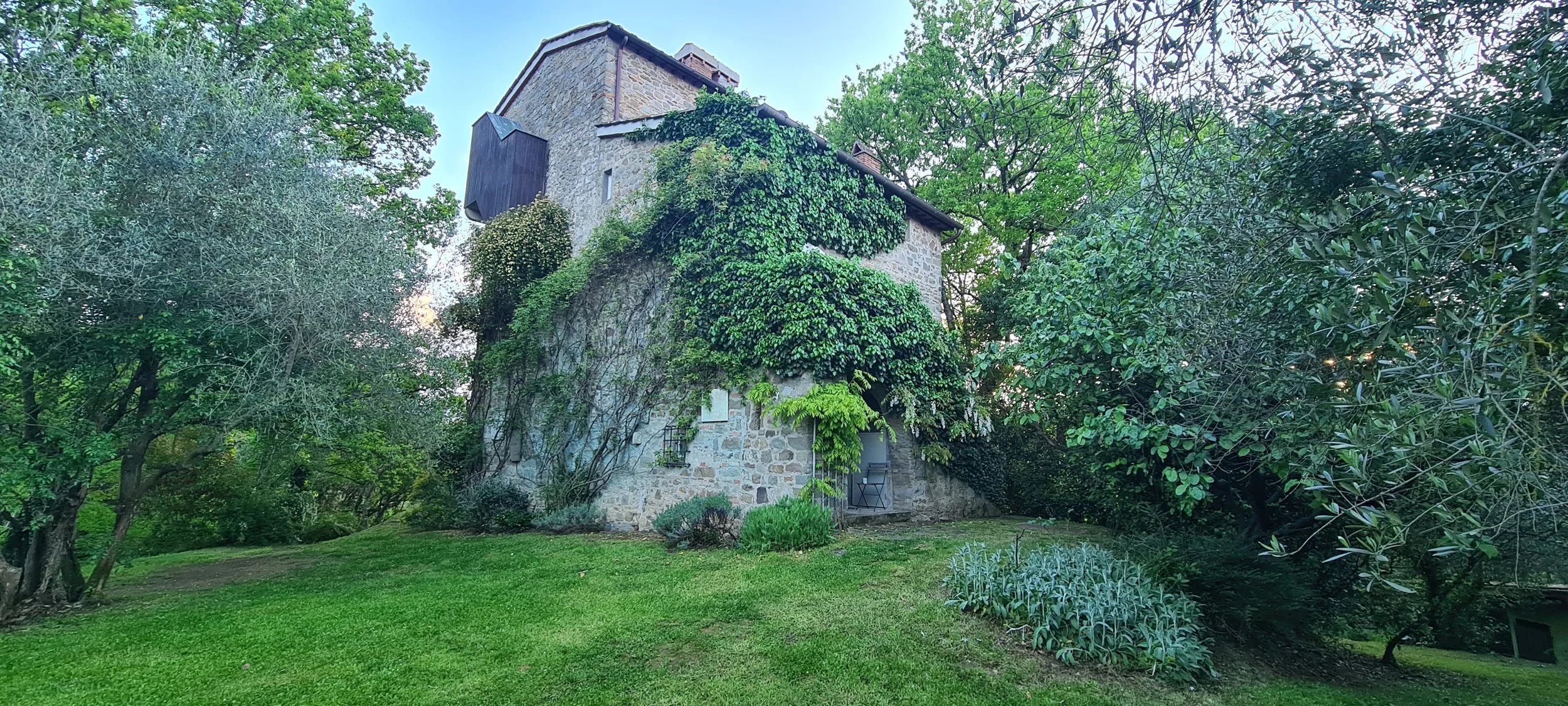 Maison en pierre entourée d'arbres et de végétation, avec une pelouse verte en premier plan.