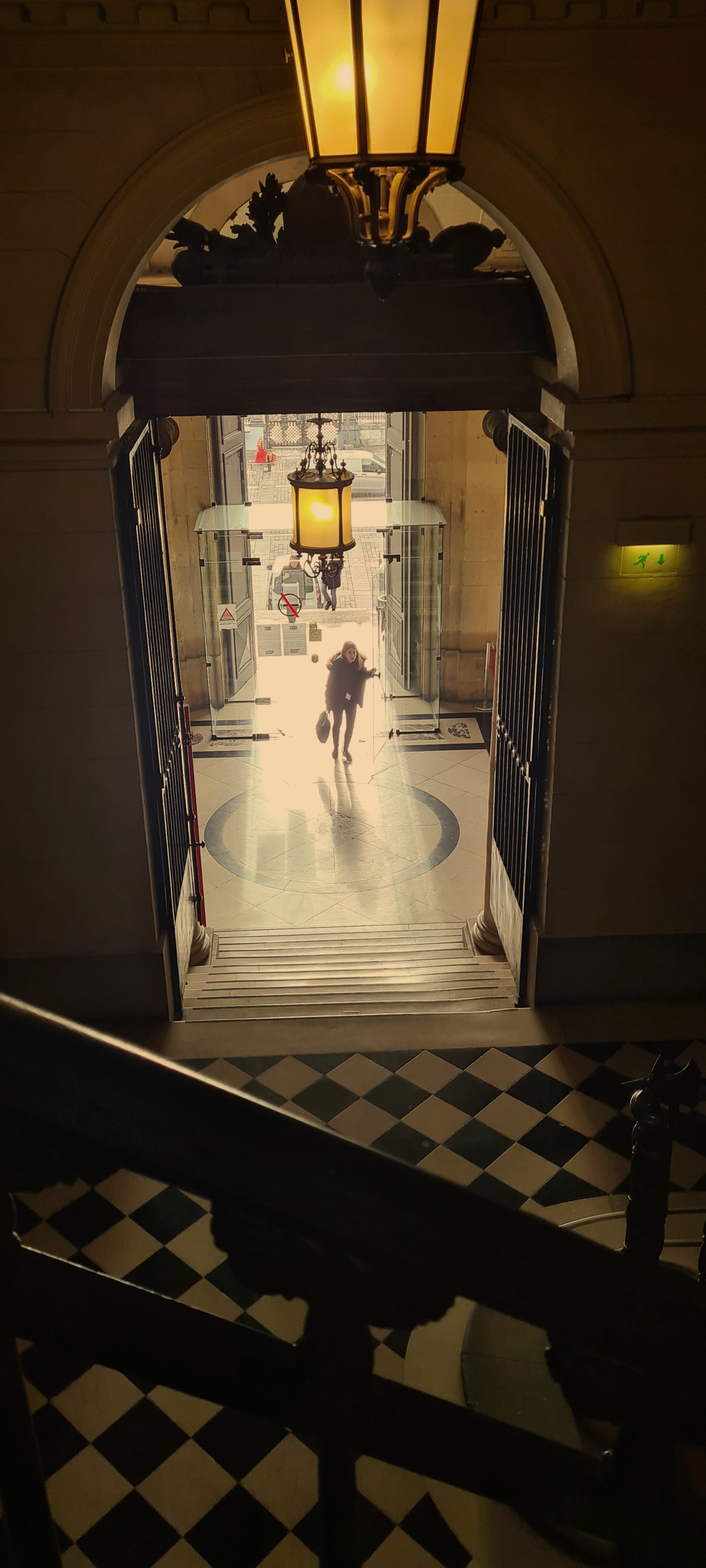 Vue intérieure d'une entrée d'immeuble avec un grand lampadaire suspendu, portes vitrées ouvertes laissant passer une personne avec un sac à la main, sol en mosaïque noir et blanc, et escalier en premier plan.