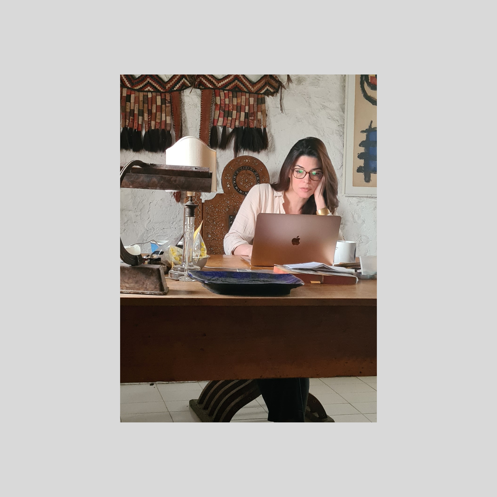 Femme travaillant sur un ordinateur portable à un bureau en bois, avec une lampe, une tasse blanche, et des papiers, dans une pièce décorée avec des œuvres d'art et un textile ethnique en arrière-plan.