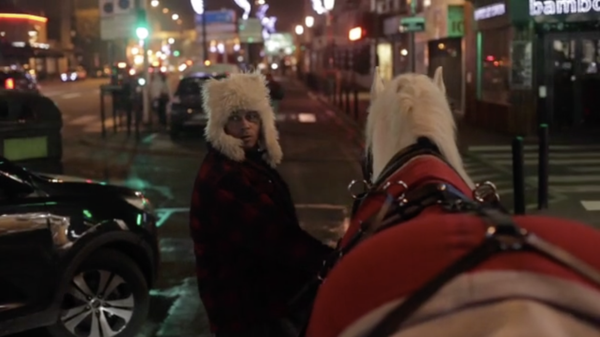 Une personne portant un bonnet en fourrure blanche est assise devant un cheval blanc, tous deux sur un trottoir dans une ville éclairée la nuit.