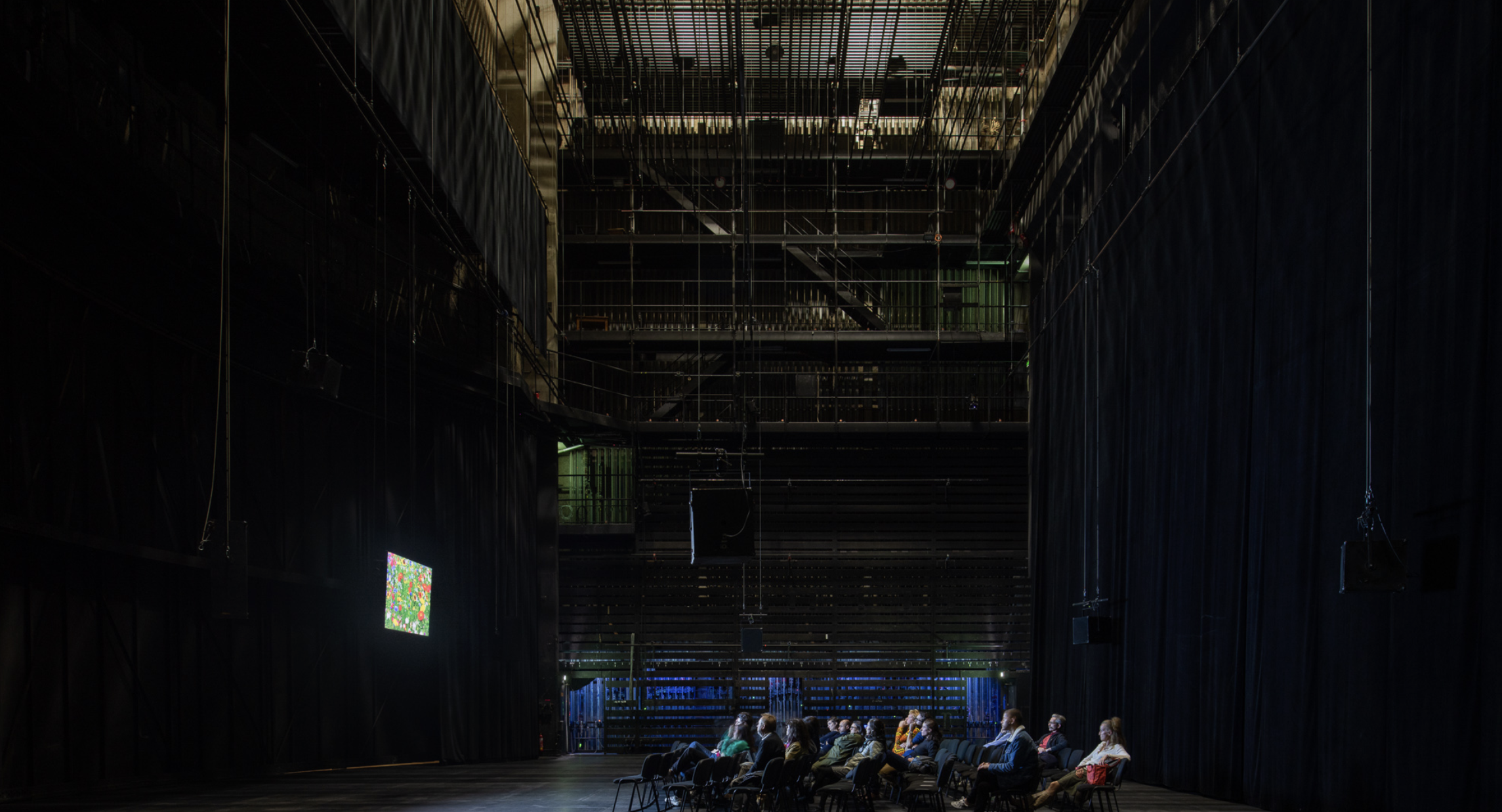 Salle de théâtre ou auditorium sombre avec quelques personnes assises, et un écran multicolore sur le mur à gauche.