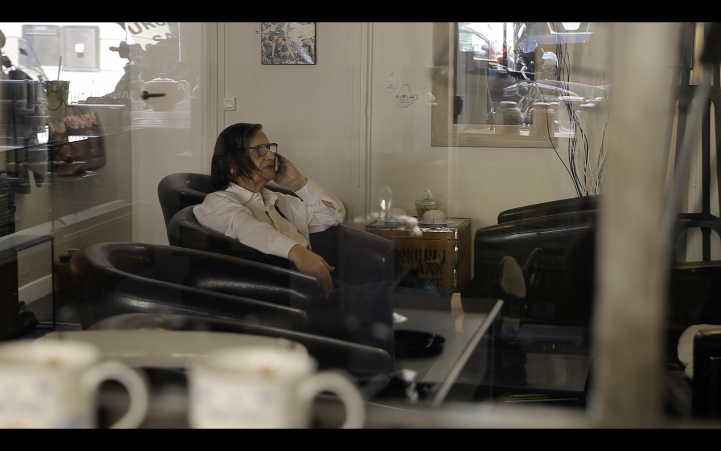Une femme assise en fauteuil, parlant au téléphone, vue à travers une vitre d'une salle de café ou de bureau.