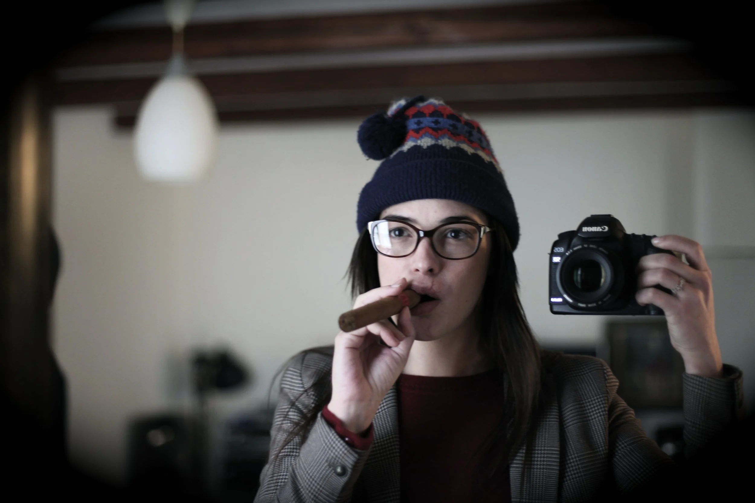Une personne prenant un selfie dans un miroir, portant un bonnet coloré, des lunettes et tenant un cigare.