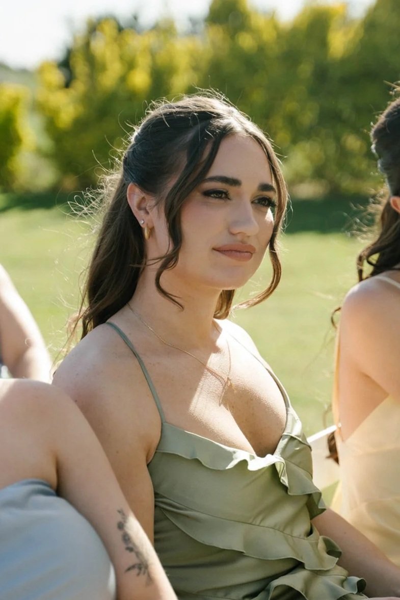 A young woman with dark hair and fair skin, wearing a green dress with ruffles, sitting outdoors in a park with green trees in the background, during daytime.