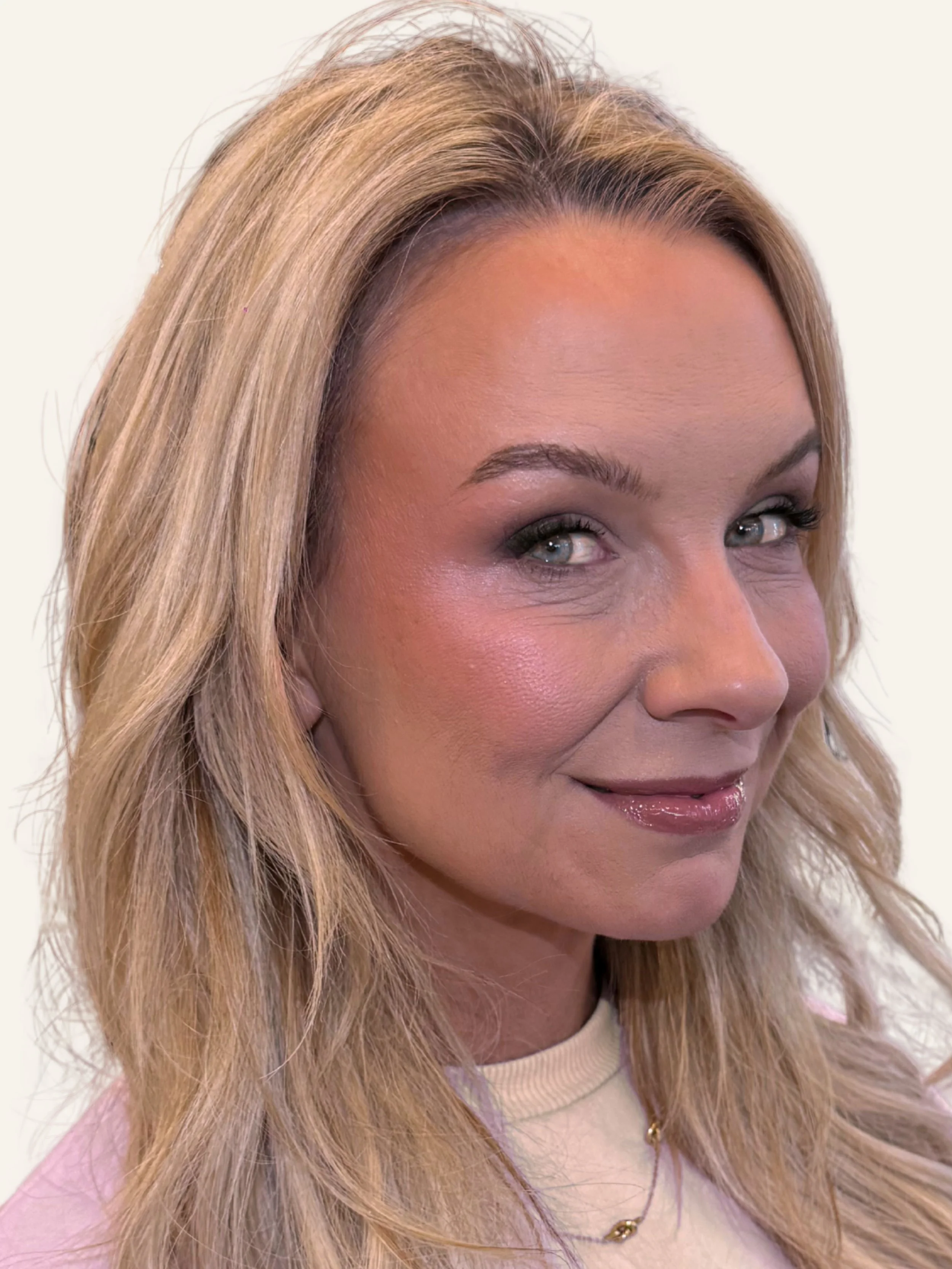 Close-up of a woman with long blonde hair and light makeup, smiling slightly. She is wearing a beige top and a gold necklace.