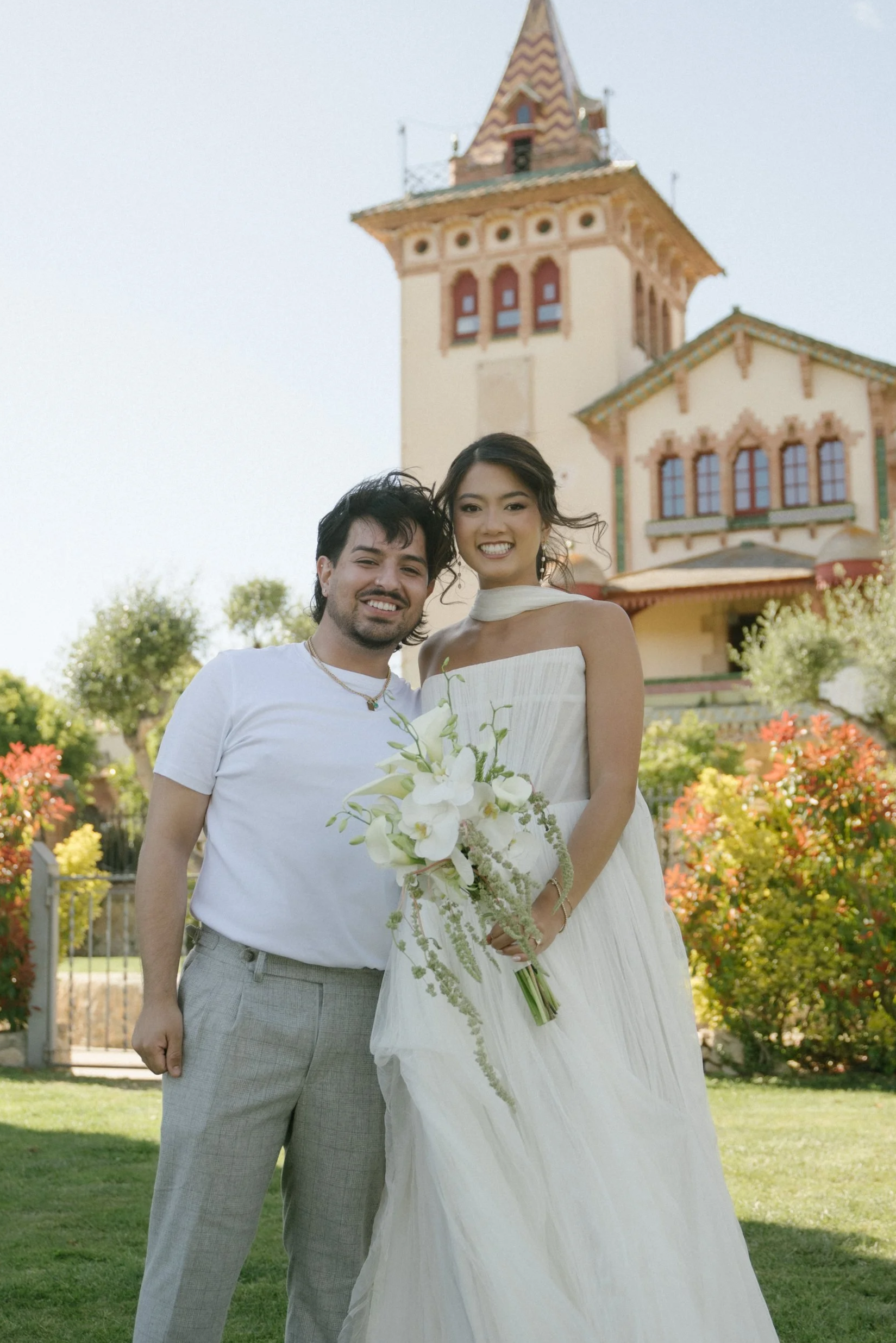 A couple stands outdoors in front of a castle-like building, smiling. The woman is wearing a white dress and holding a bouquet, and the man is dressed in a white t-shirt and light-colored pants.