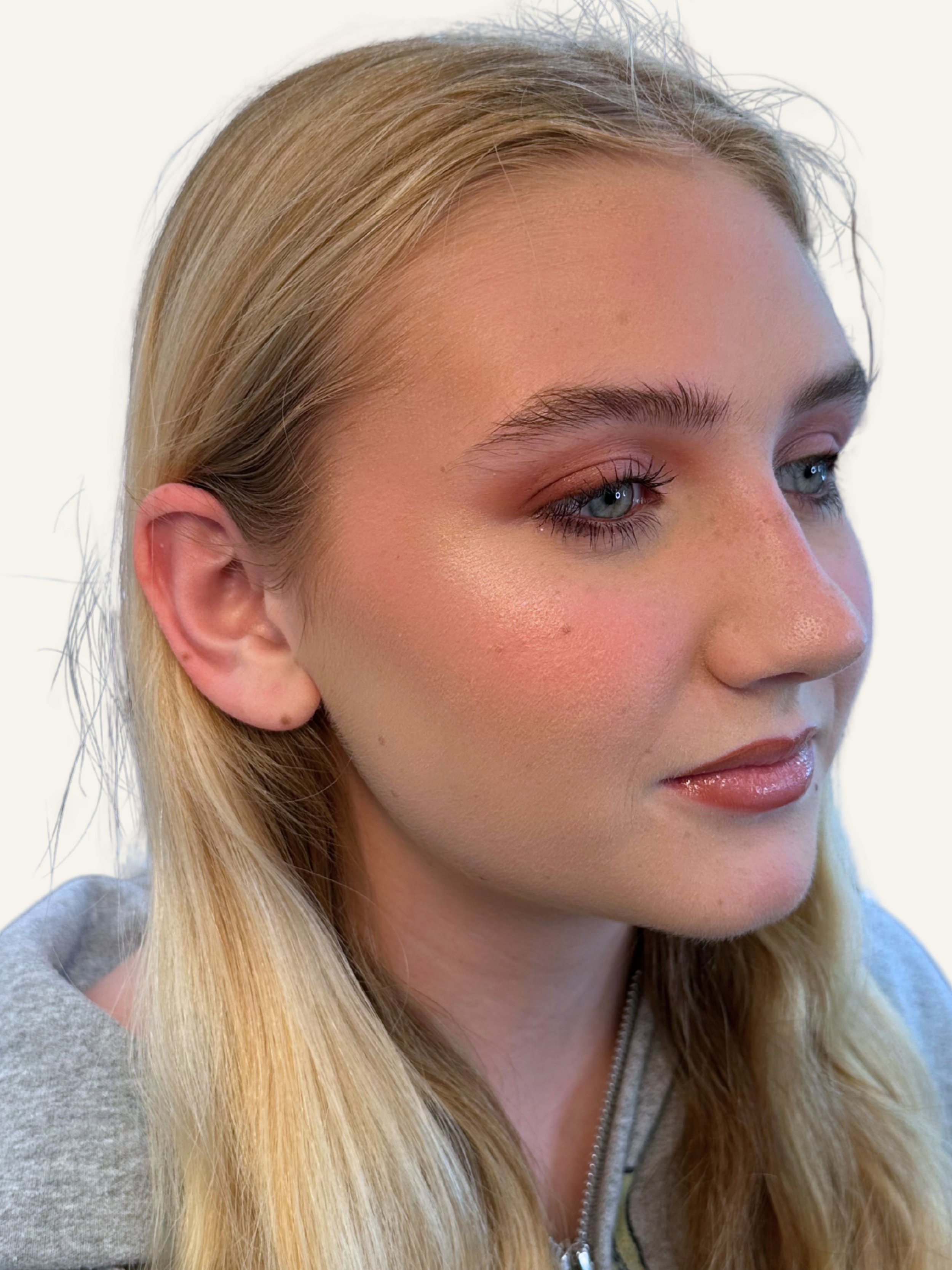 Close-up of a woman with blonde hair, blue eyes, light makeup, pink blush, and shiny lips, wearing a grey top.
