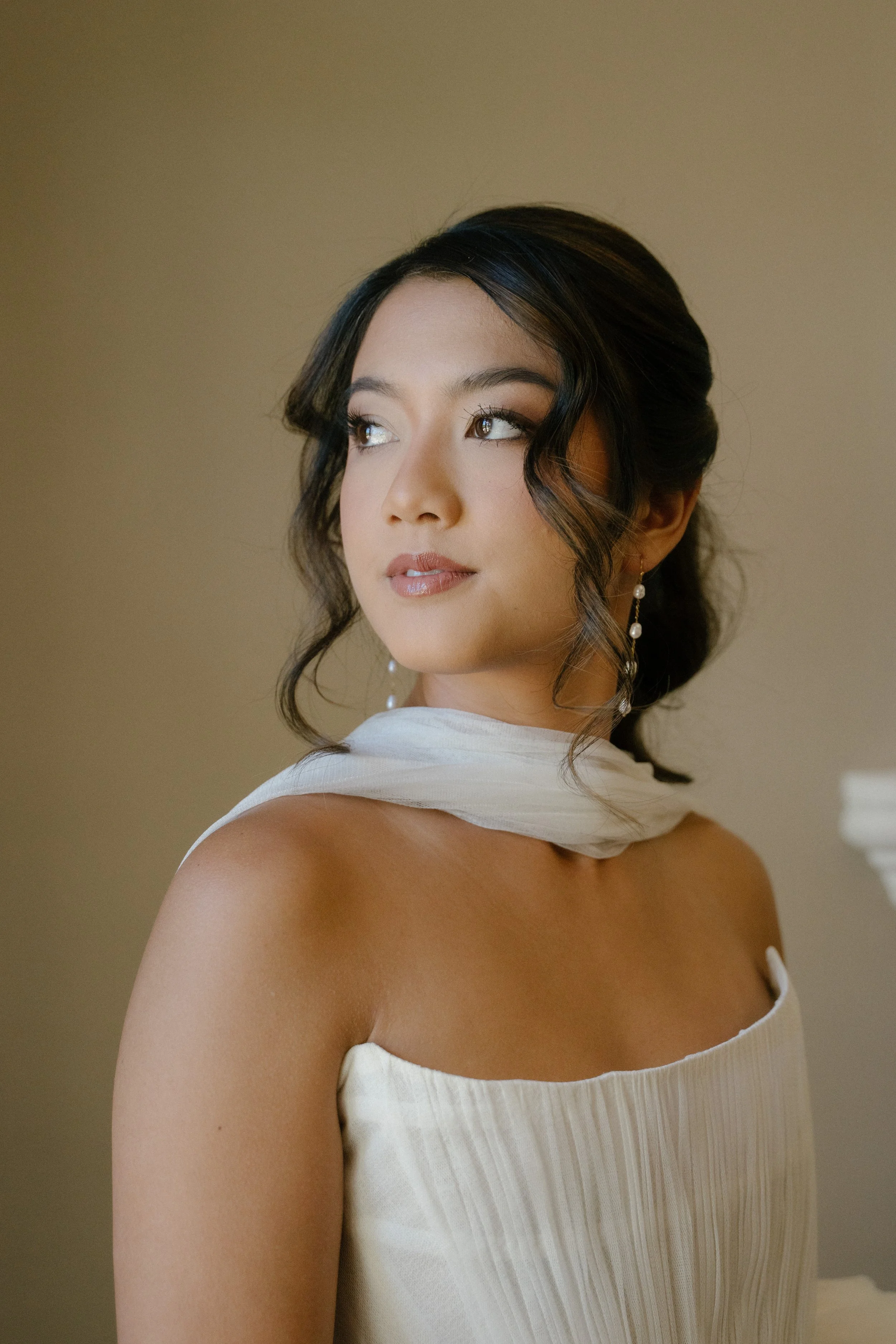 A woman with dark hair styled in waves, wearing pearl earrings, a white strapless dress, and a white scarf around her neck, looking to the side with a soft expression, against a neutral background.