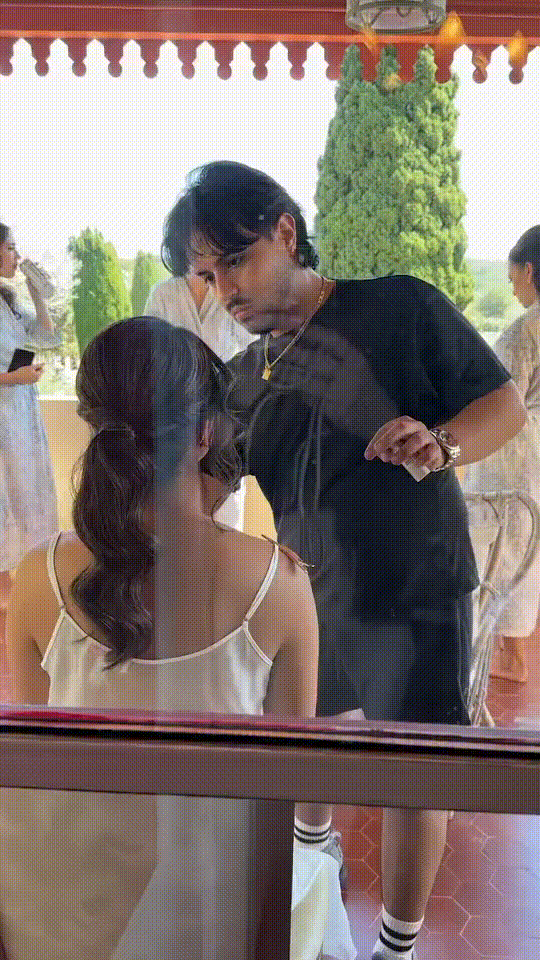 A man with dark hair applying makeup or face paint to a woman sitting outside near a window, with trees and other people visible in the background.