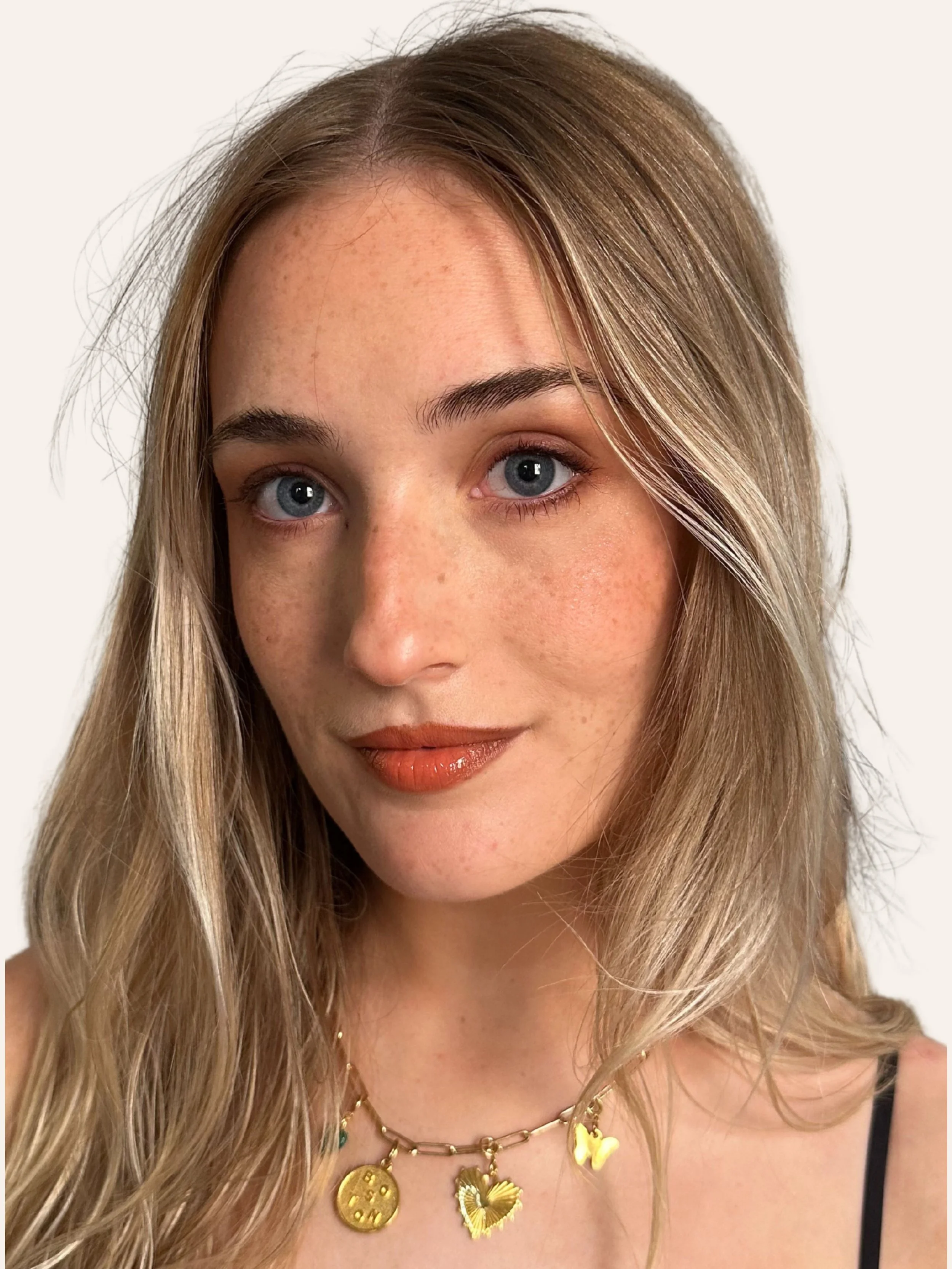 Close-up portrait of a young woman with blue eyes, light brown hair, and freckles, wearing a gold necklace with charms, a black strap top, and natural makeup.