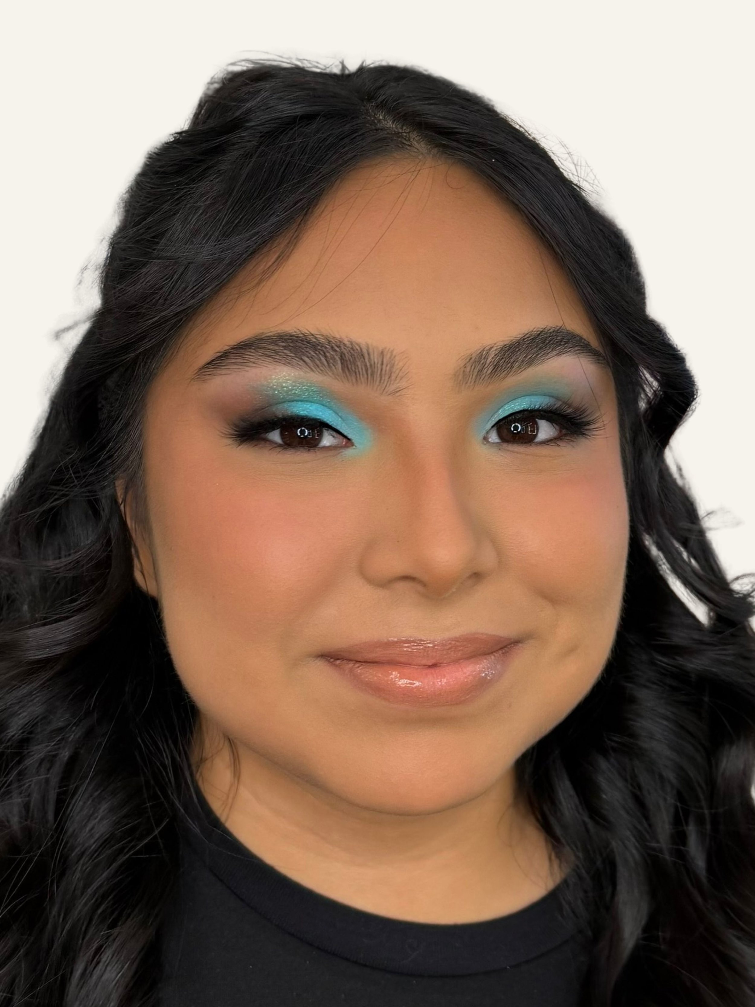 Close-up of a woman with long black wavy hair and makeup featuring blue eyeshadow, dark eyeliner, and glossy lips.