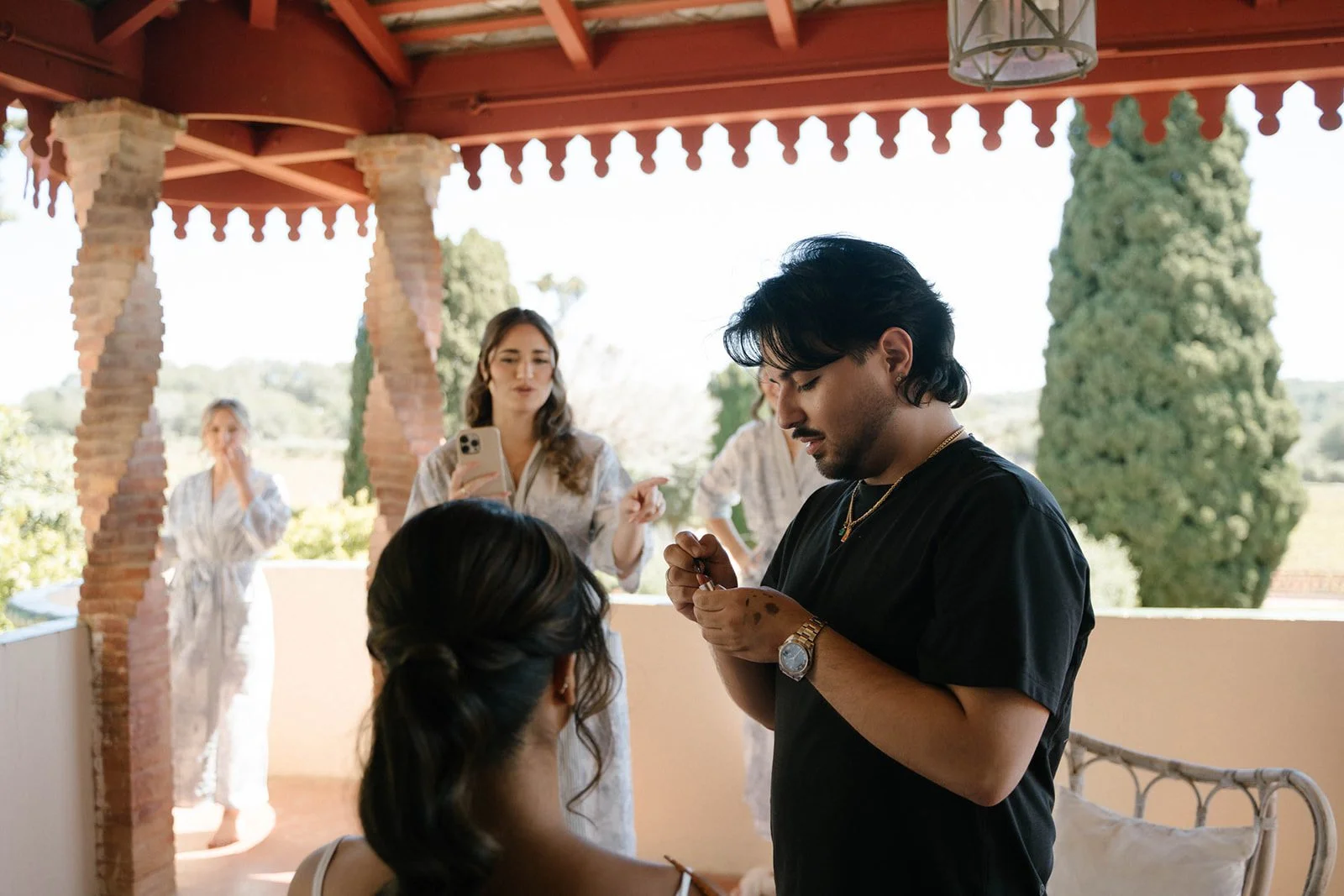 A man in black shirt applying makeup to a woman seated on a cushioned outdoor porch, with three women in the background dressed in robe-like garments. The setting features a wooden roof, brick columns, and greenery outside.
