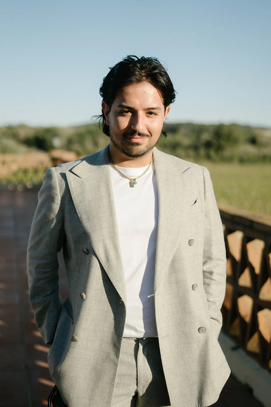 A young man with dark hair and a beard, dressed in a light gray double-breasted suit and white T-shirt, standing outdoors on a patio with greenery in the background.