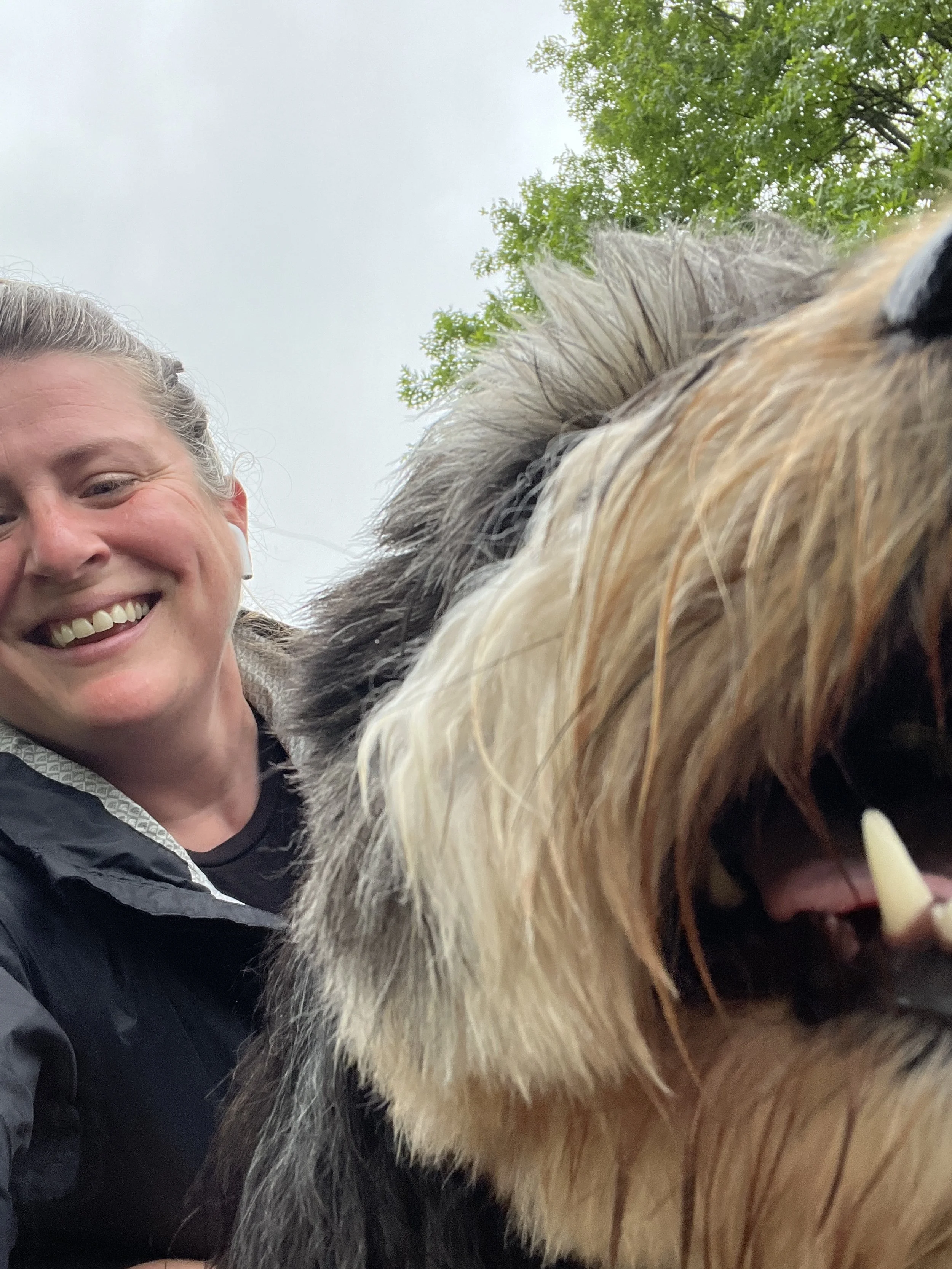 A smiling woman taking a close-up selfie with a large, fluffy dog outdoors under cloudy skies.