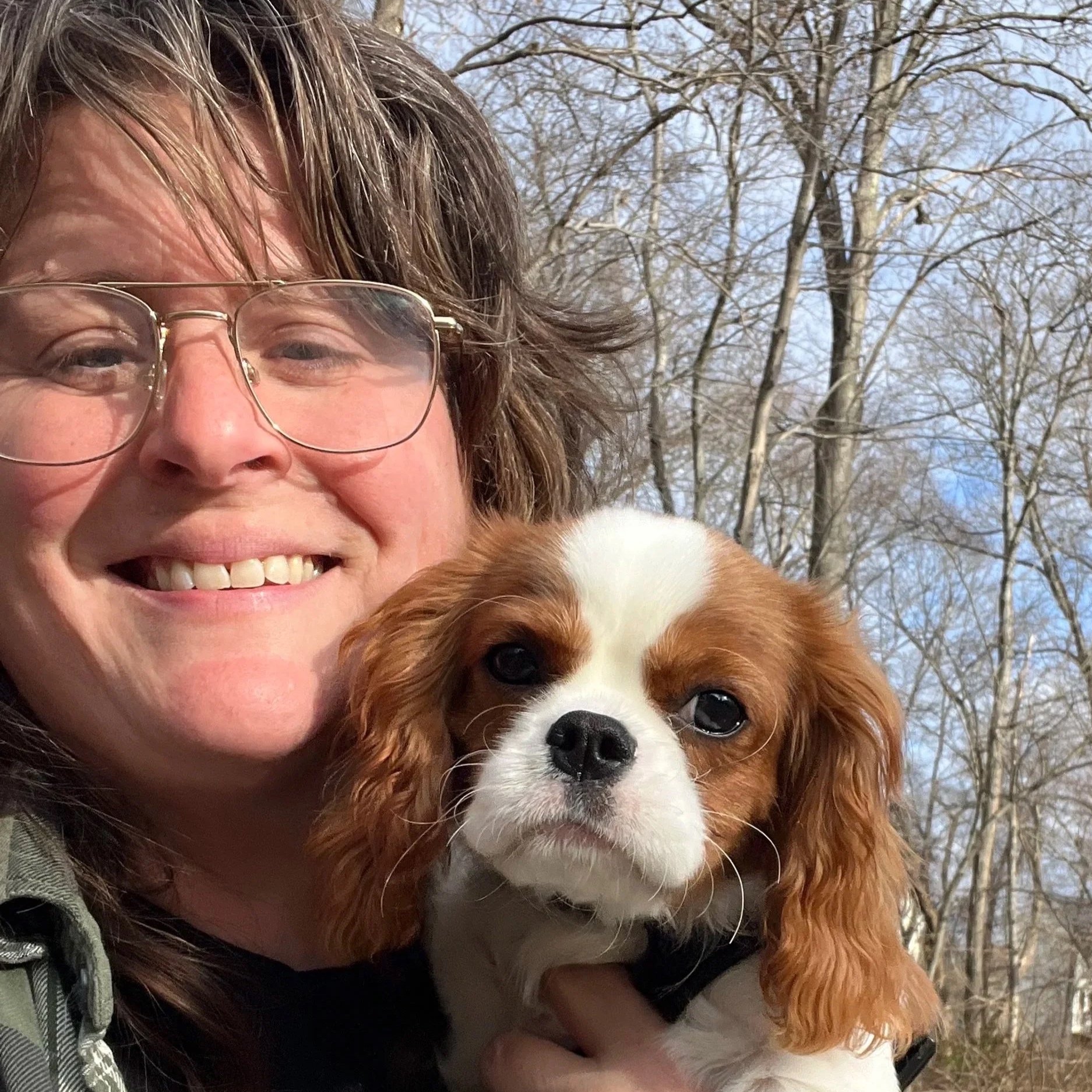 A smiling woman with glasses holding a Cavalier King Charles Spaniel outside with leafless trees and a blue sky in the background.