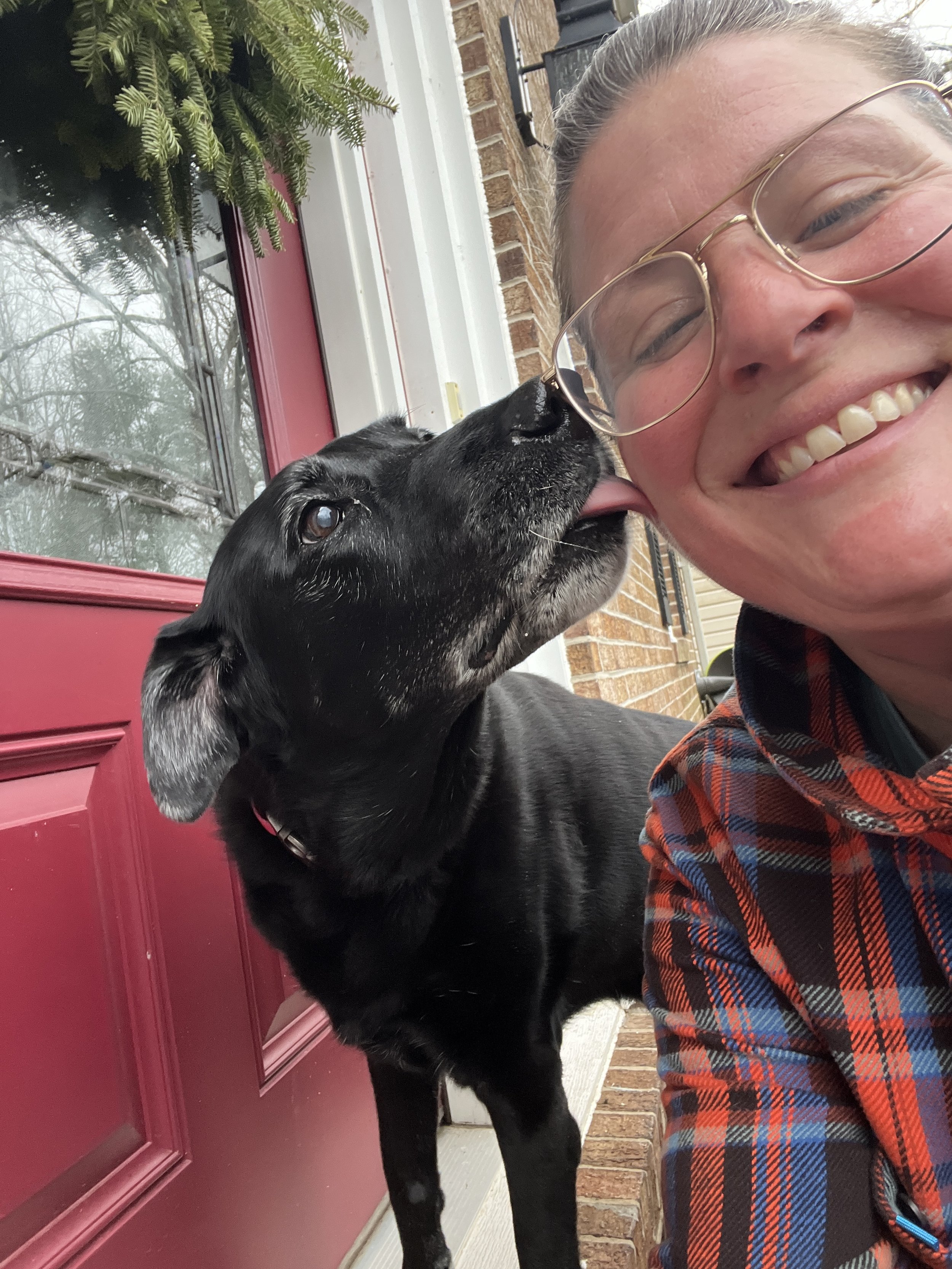 A woman with glasses smiling as a black dog licks her cheek outside a house with a red door and brick wall.