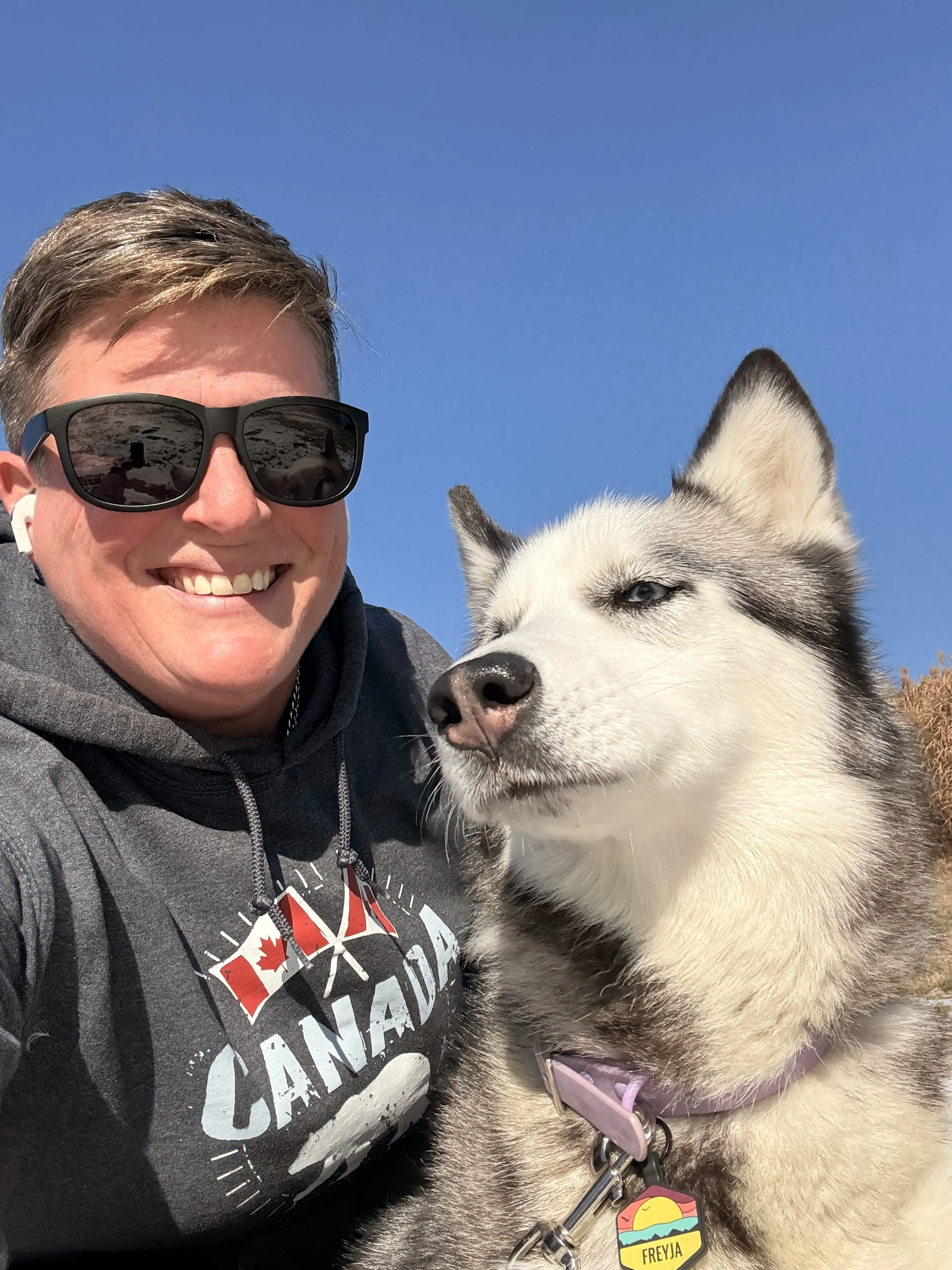 A smiling person with sunglasses and a gray hoodie taking a selfie outdoors with a husky dog against a clear blue sky.