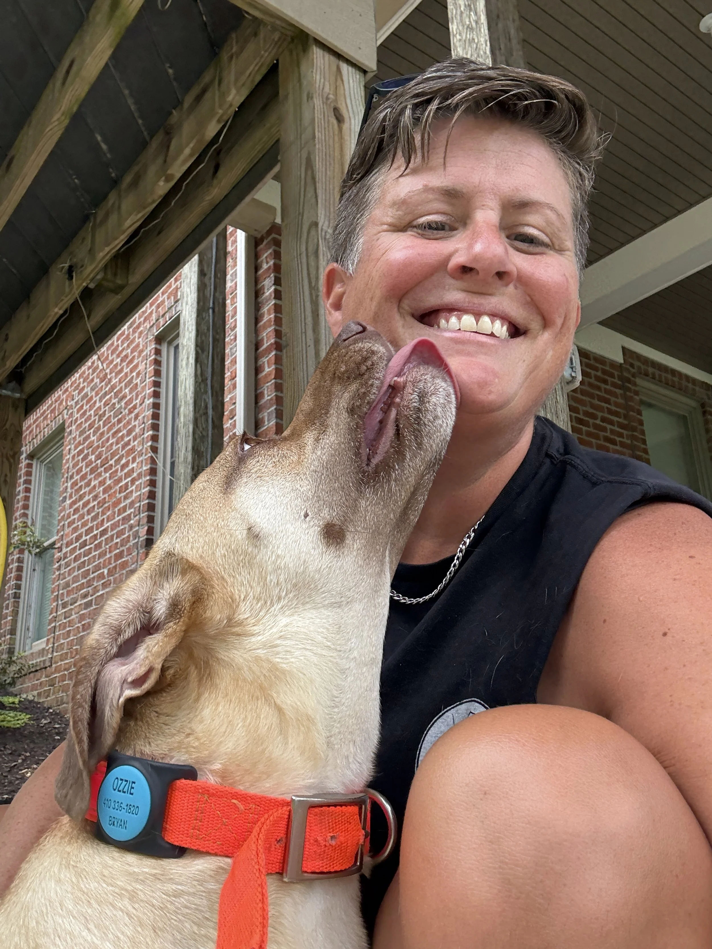A smiling woman with short, gray hair wearing a black sleeveless shirt, taking a selfie with a light-colored dog with a red collar. The dog is touching the woman's face with its nose and tongue. They are outdoors near a brick house and wooden porch.