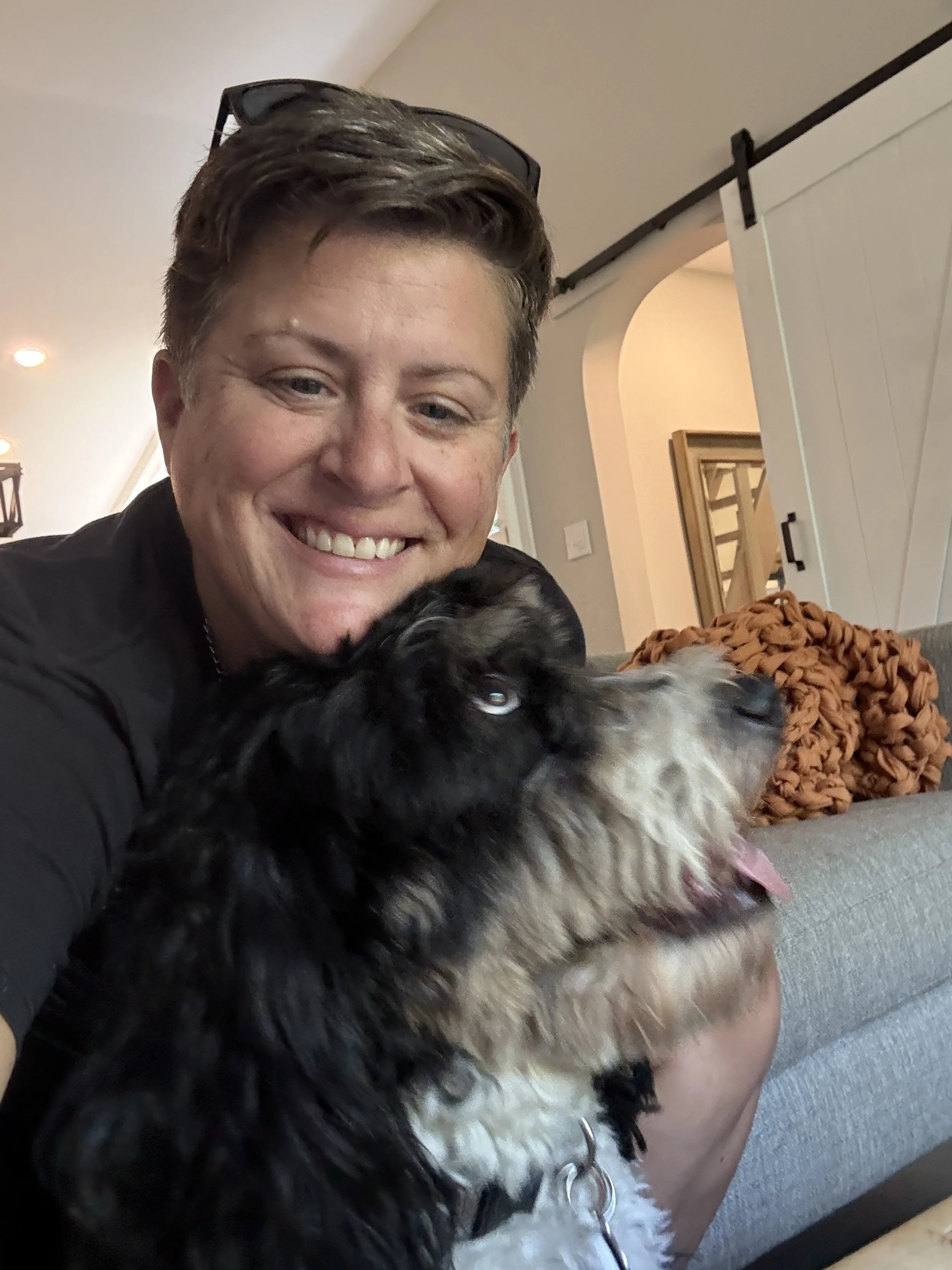 A person smiling and hugging a black and white dog on a grey couch inside a home.
