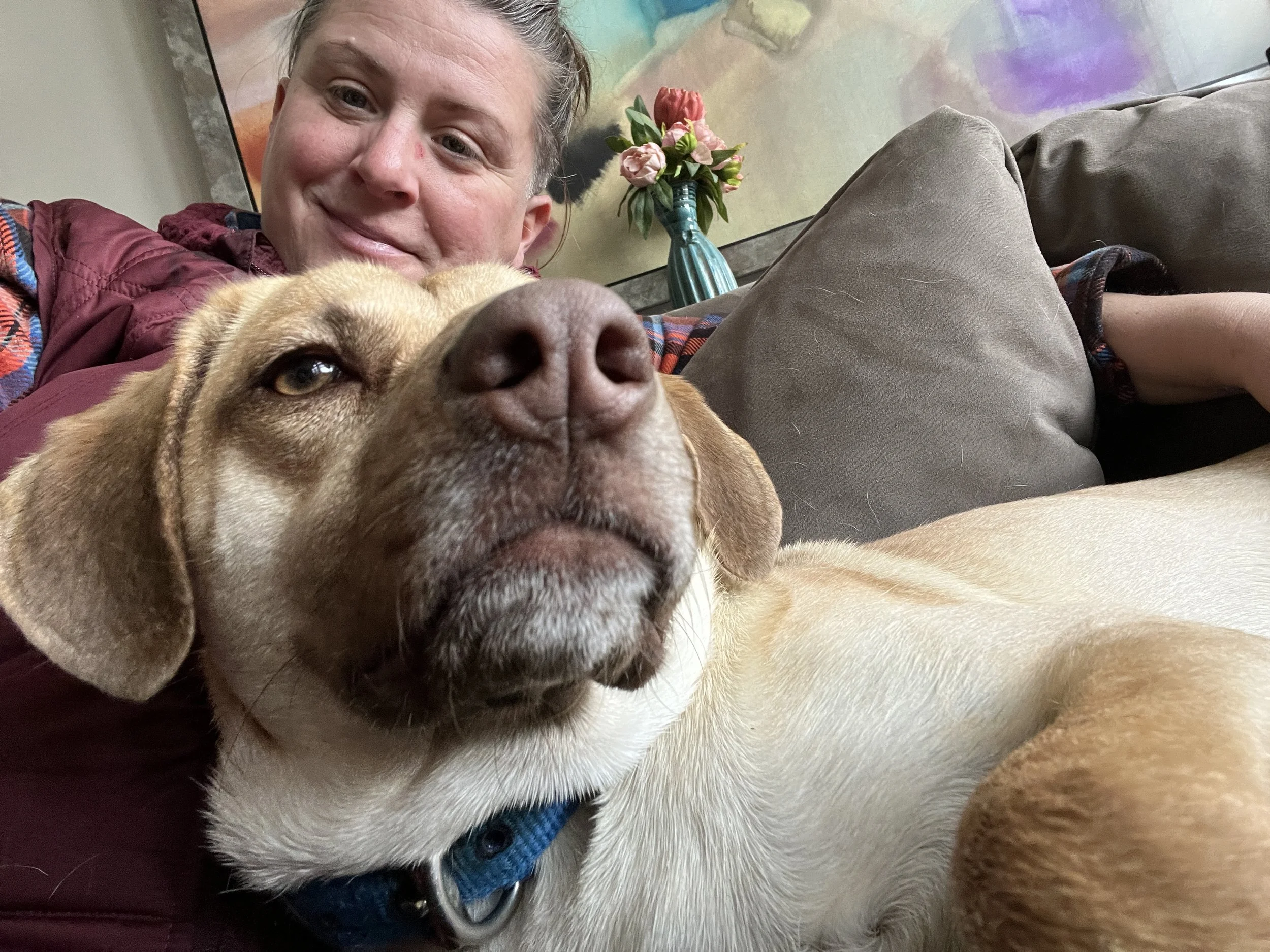 A woman and a large tan dog are lying on a couch. The woman is smiling, and the dog is relaxed with one eye partially closed. The background shows a colorful abstract painting and a small bouquet of pink flowers in a teal vase.