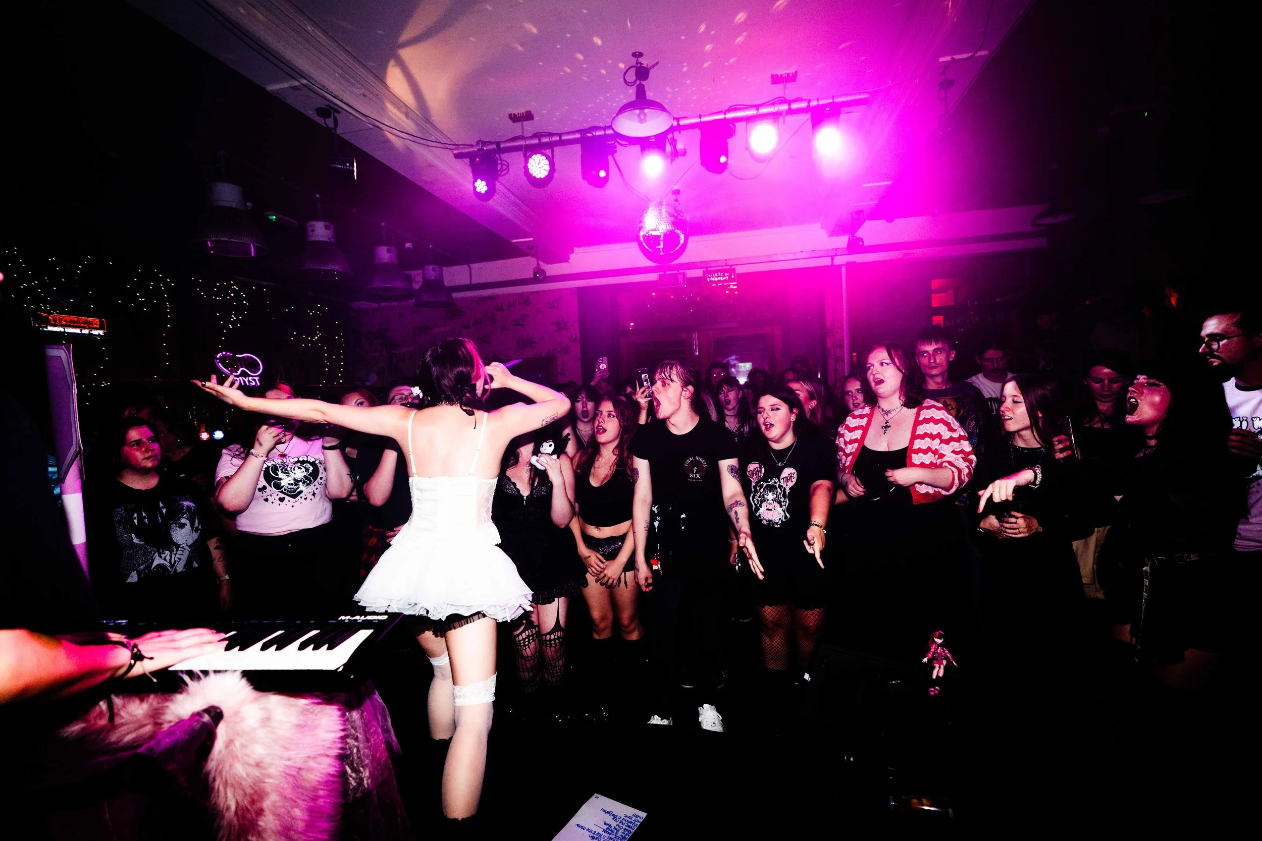 A live music event with a singer in a white dress performing for an audience at a nightclub with pink and purple lighting.