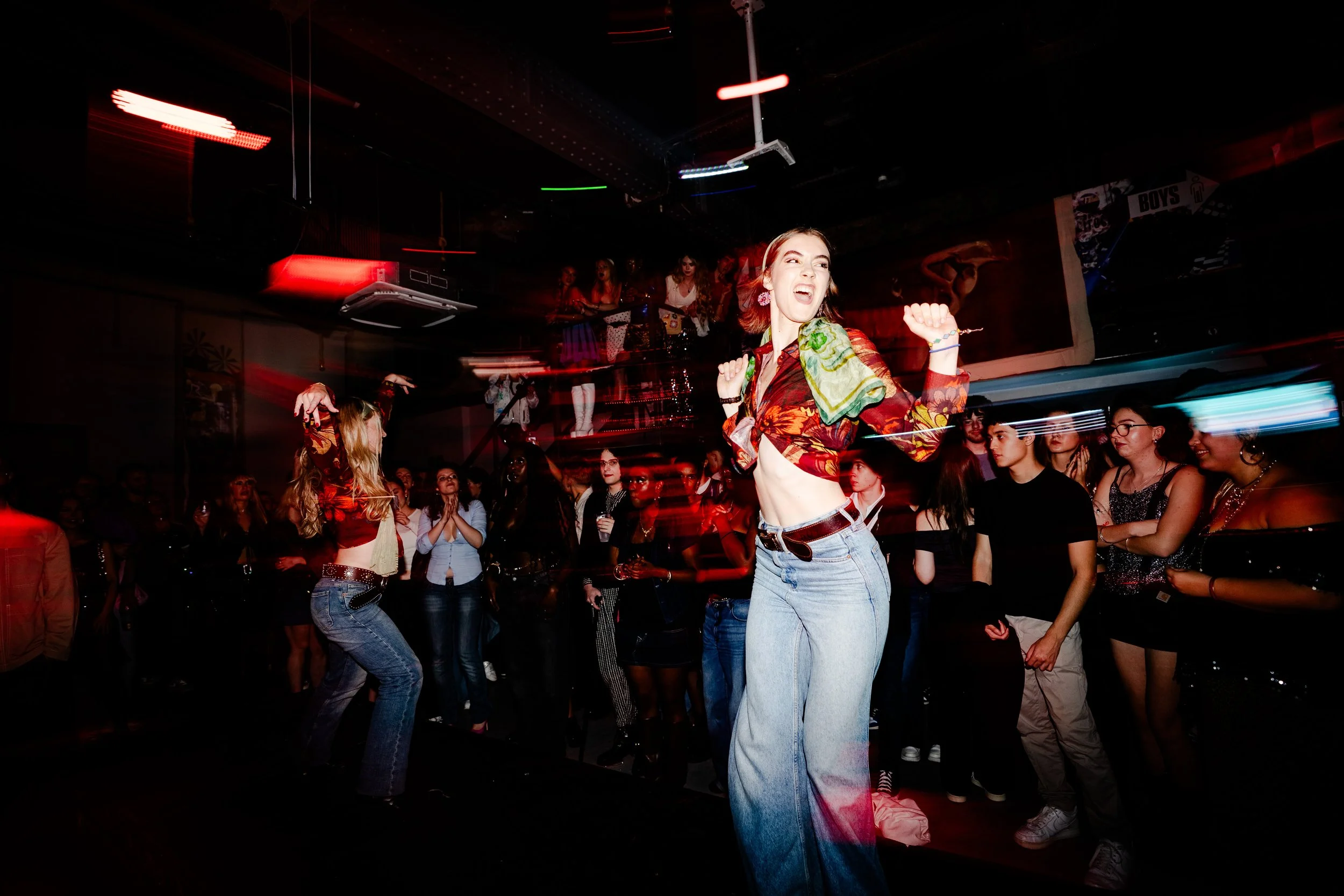 A woman dancing happily at a nightclub with other people watching her, colorful lights, and a dark interior.