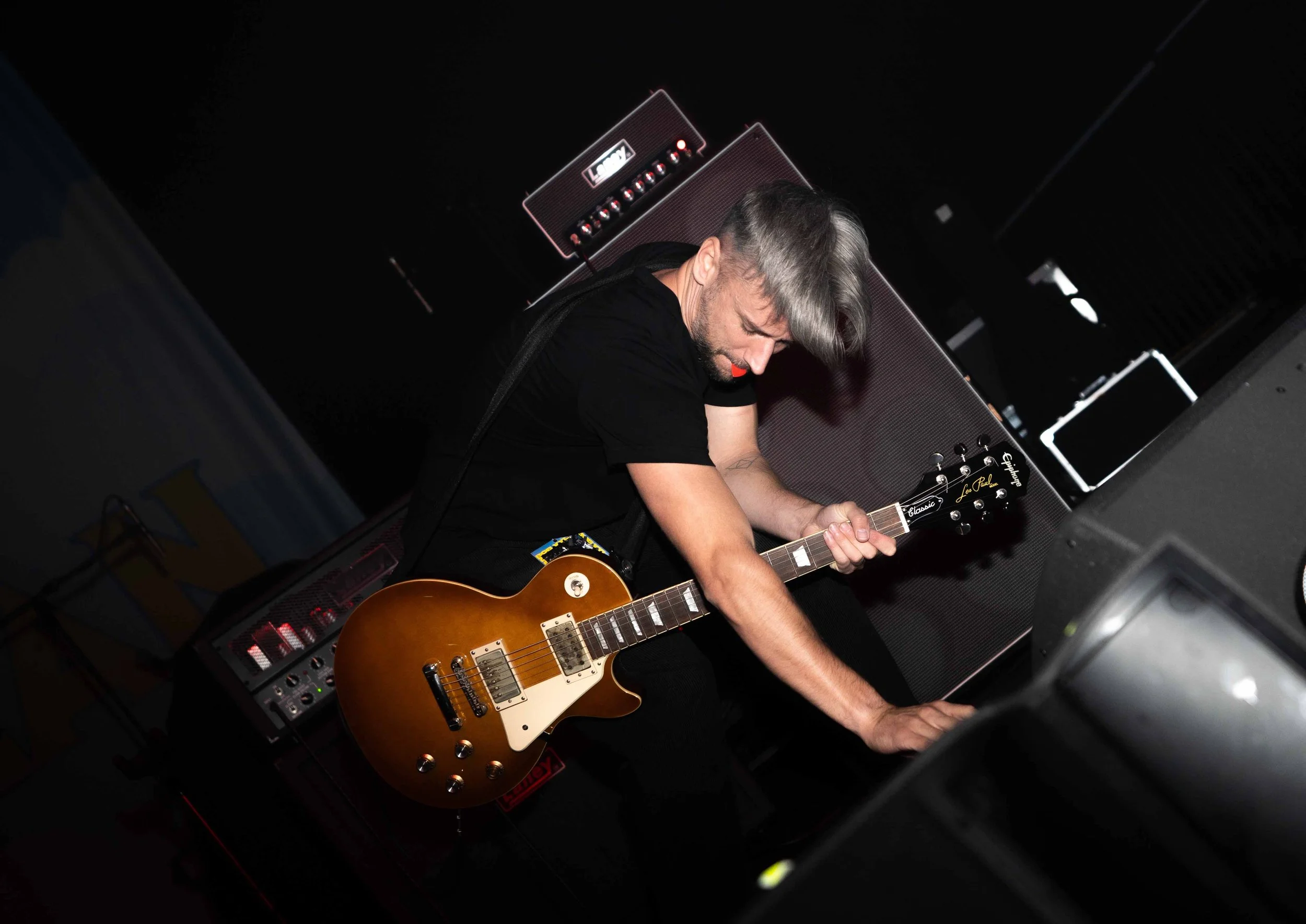 A musician with gray hair and a beard playing an electric guitar on stage, with large speakers and equipment in the background.