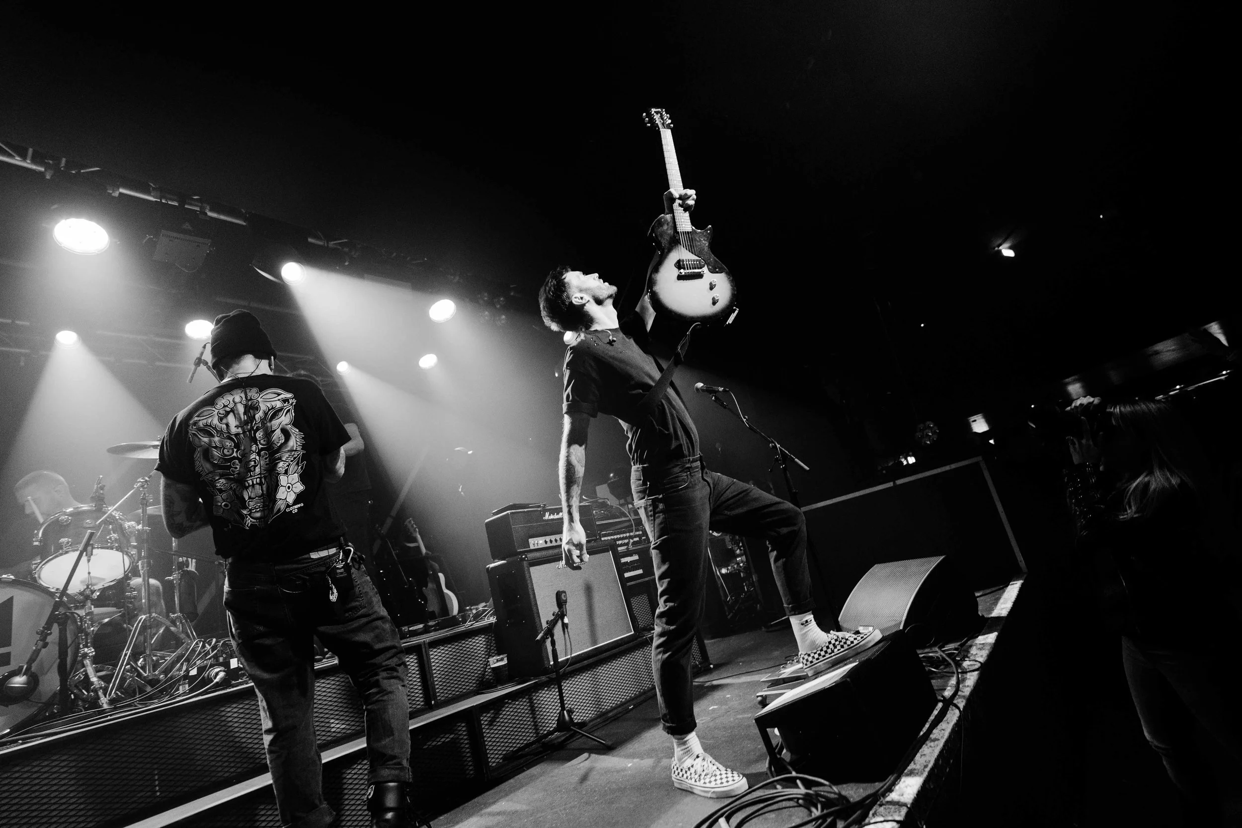 A black and white photo of a band performing on stage, featuring a guitarist raising his guitar in the air, with led lights and a drum set in the background.
