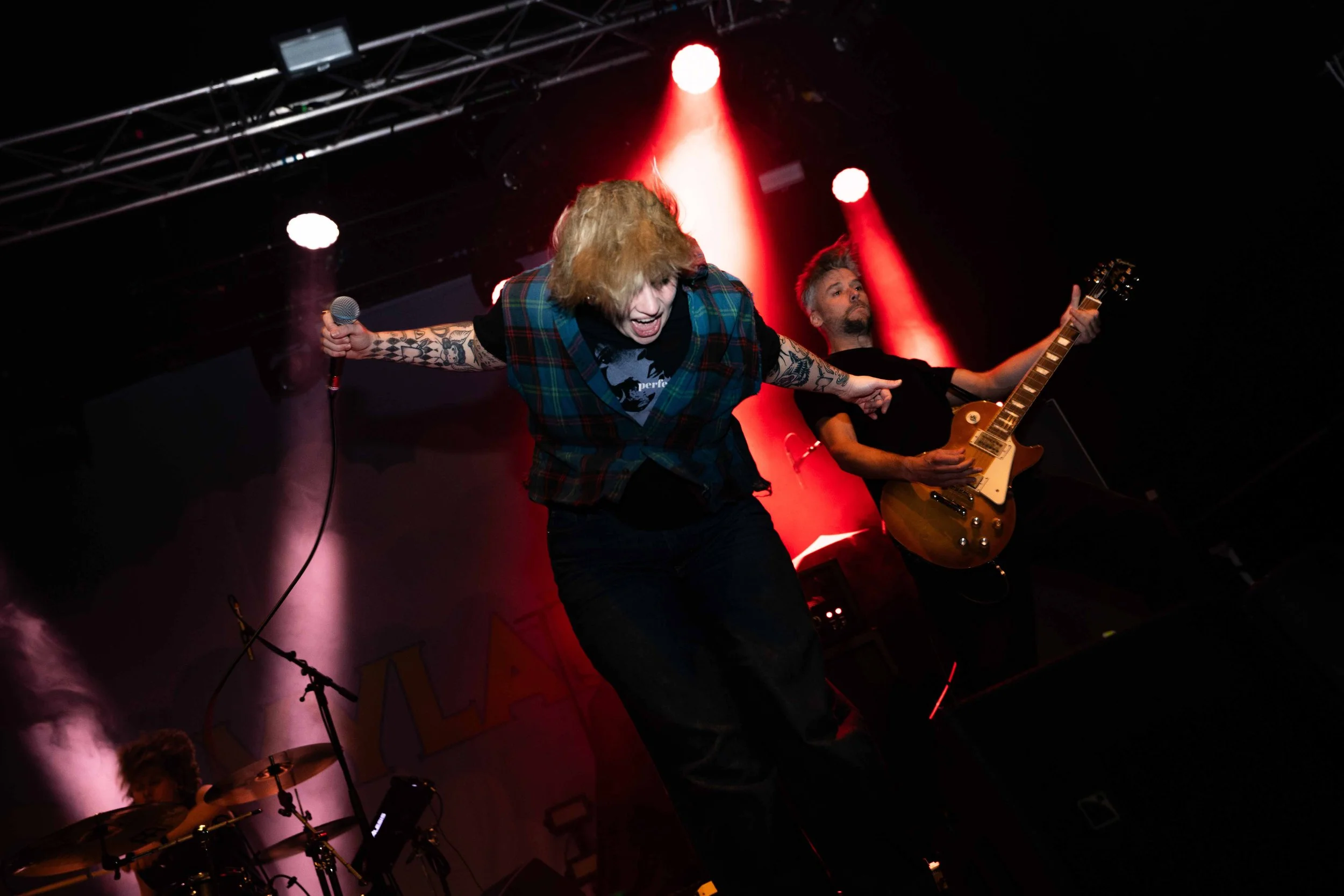 Two musicians performing on stage under red and white spotlights, with a drummer in the background. The lead singer is holding a microphone and leaning forward, with arms outstretched, wearing a plaid shirt and black pants, and the guitarist is playi