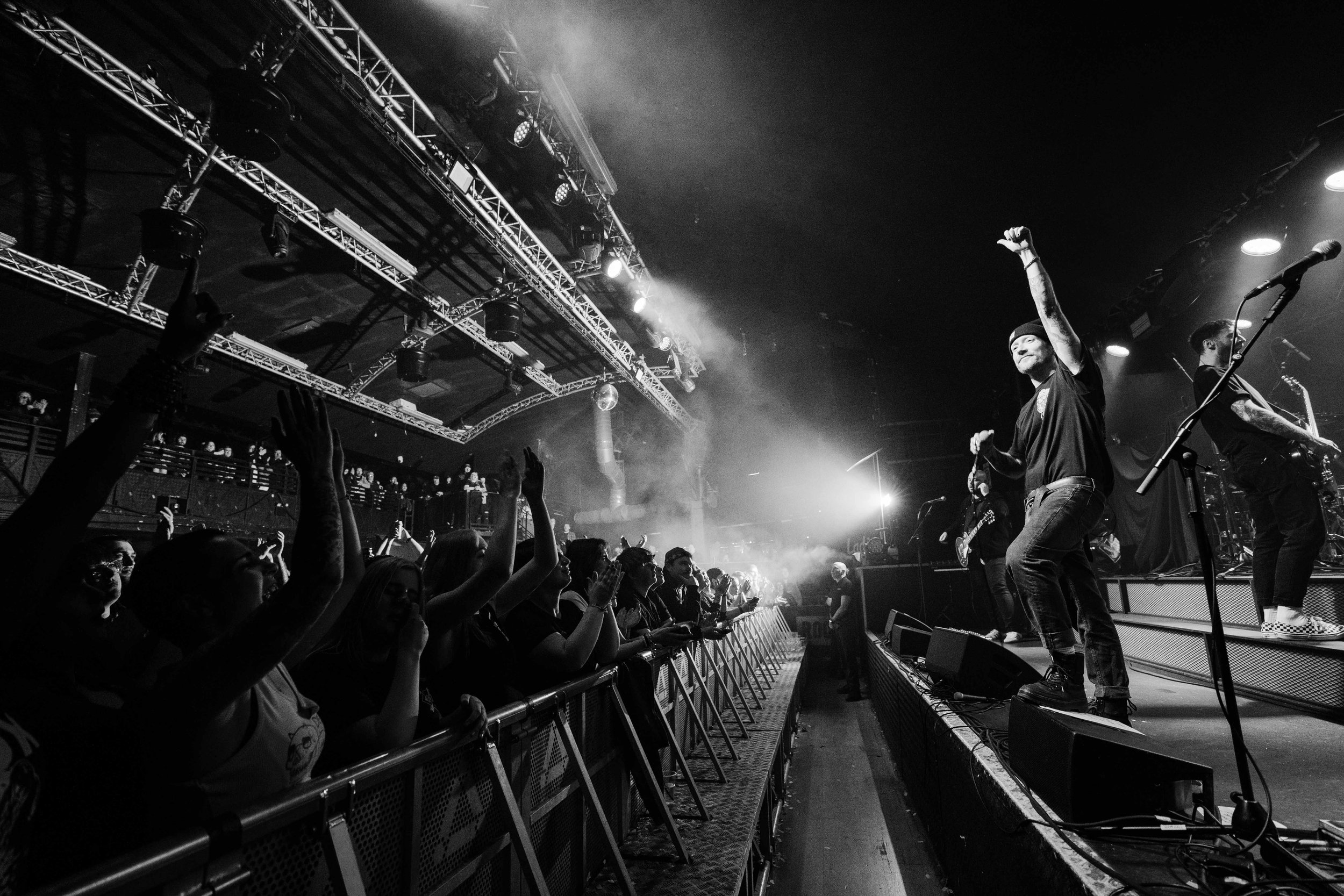 A black-and-white photo of a live concert with a band performing on stage, the lead singer with one arm raised, and an audience with their hands in the air.