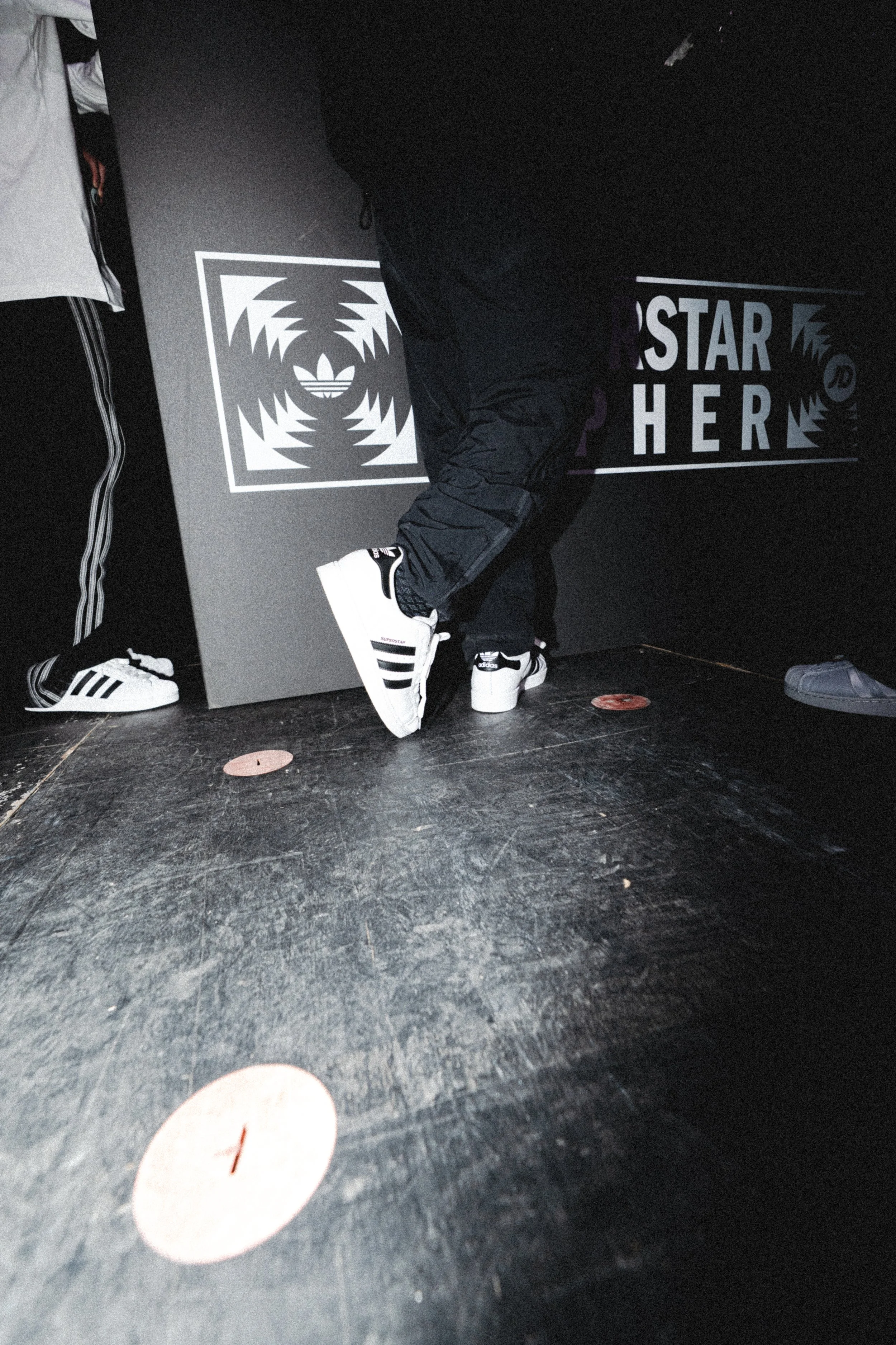 View of a dark gym or dance studio with black flooring, shoes visible, a person standing near a wall with graffiti-style art, and text that reads 'STAR HERE' on the wall.