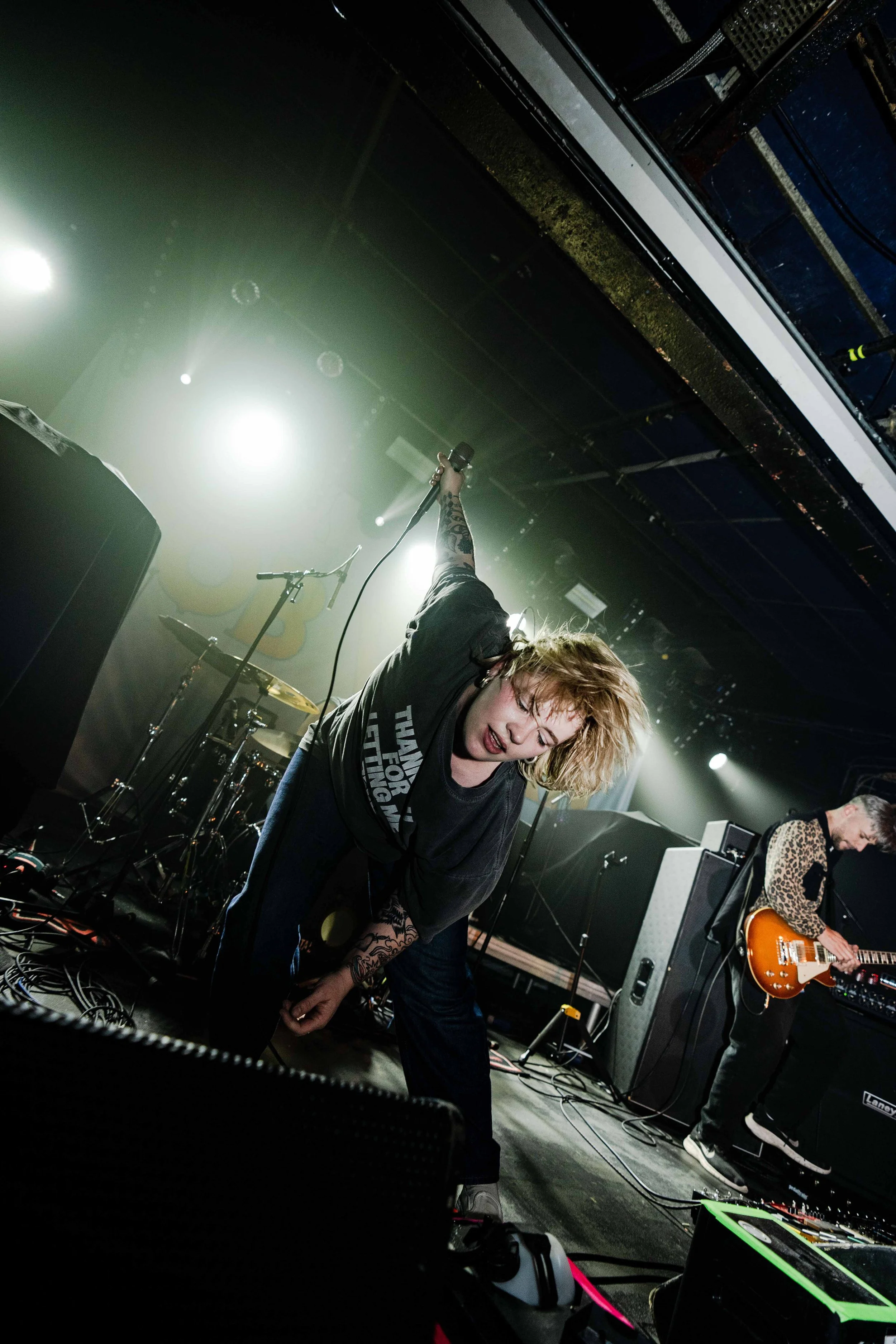 A female singer with curly blonde hair, tattoos, and a casual black t-shirt, performing passionately on stage with a microphone in hand. A guitarist in a leopard print jacket plays behind her, and stage lights create a vibrant concert atmosphere.