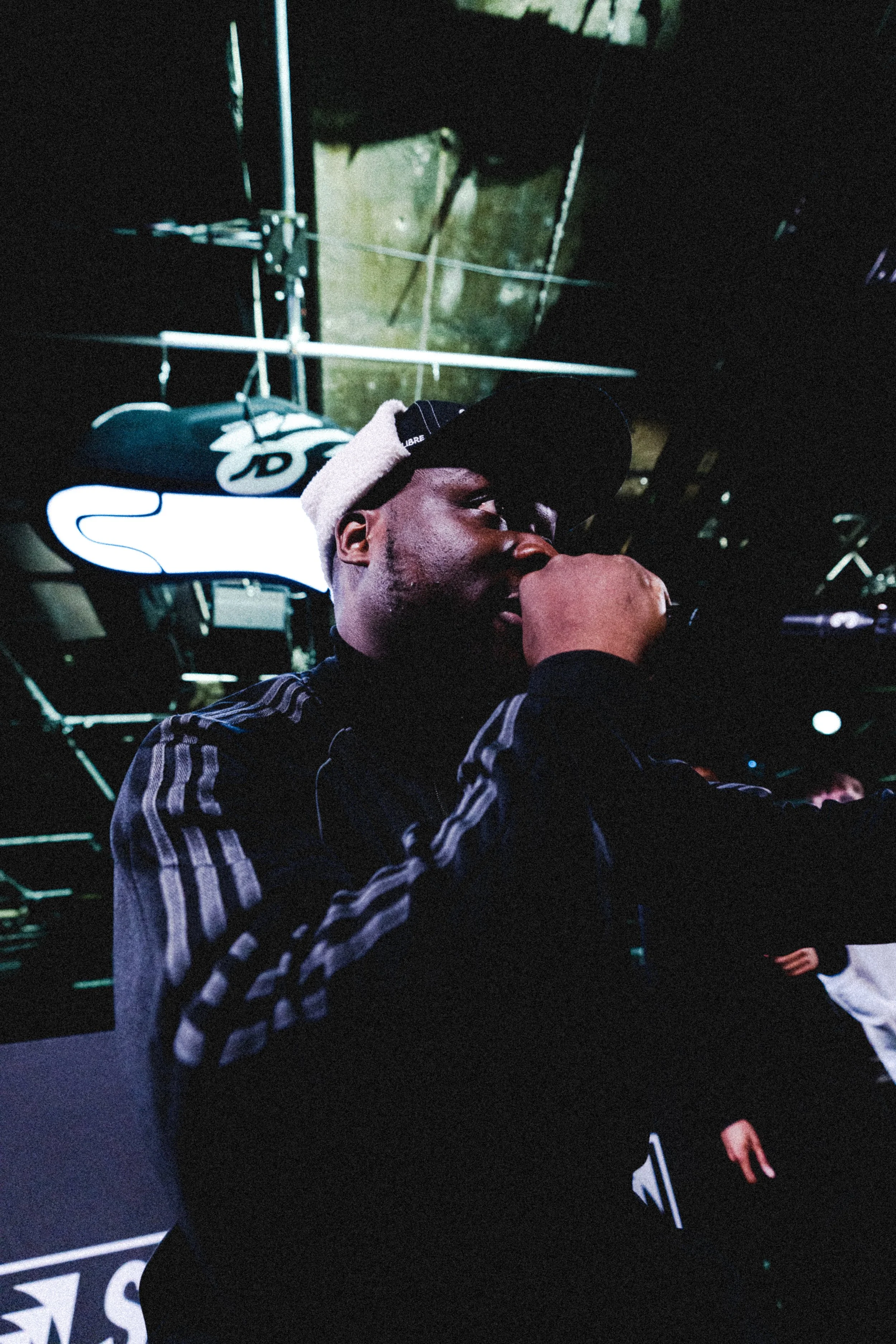 A man wearing a black cap and jacket, holding a microphone close to his mouth, performing in a dimly lit venue with a neon sign in the background.